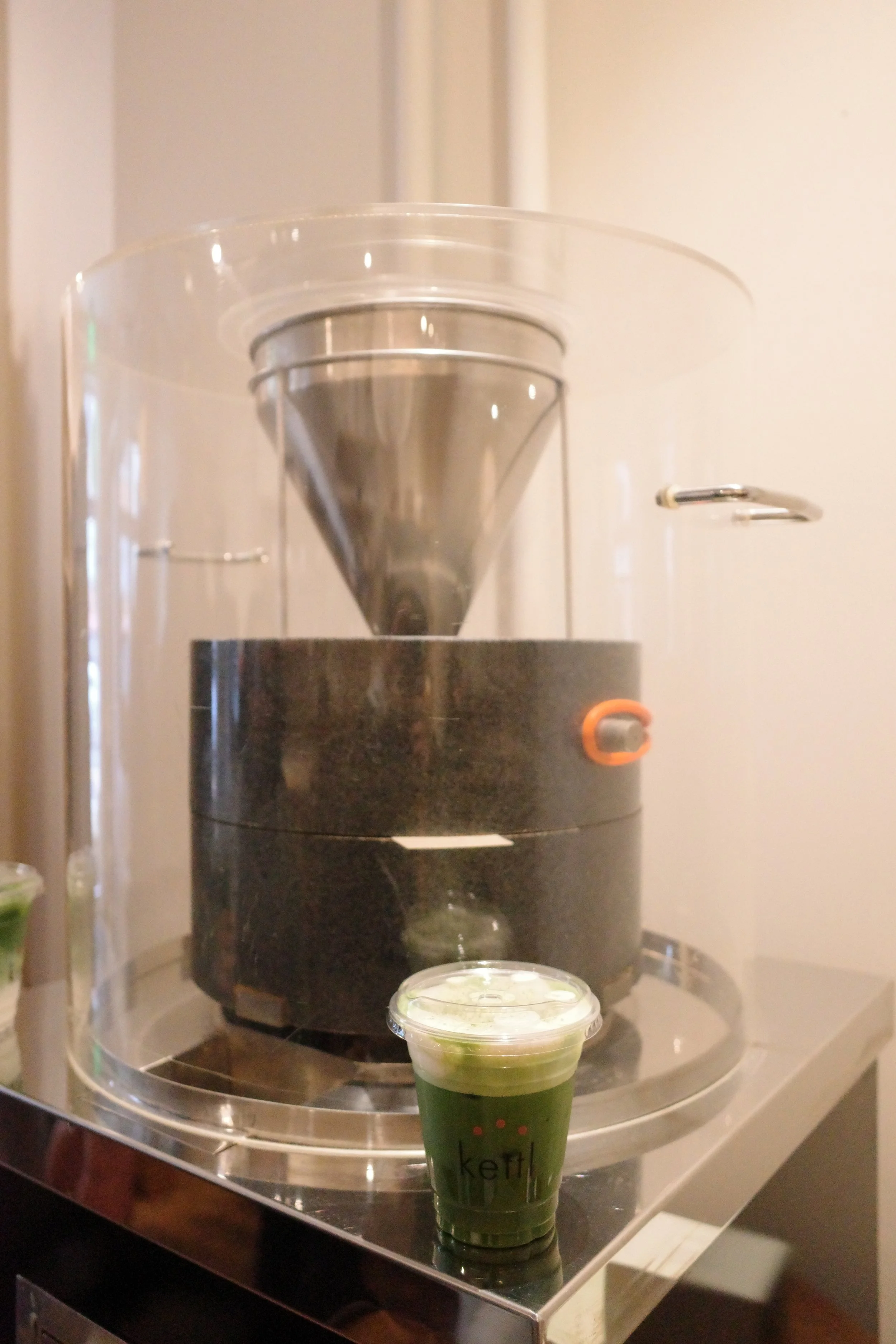 A black tea kettle with a metal cone on top, sitting on a rounded black base. A small clear plastic cup filled with a green drink with foam on top, placed in front of the kettle.