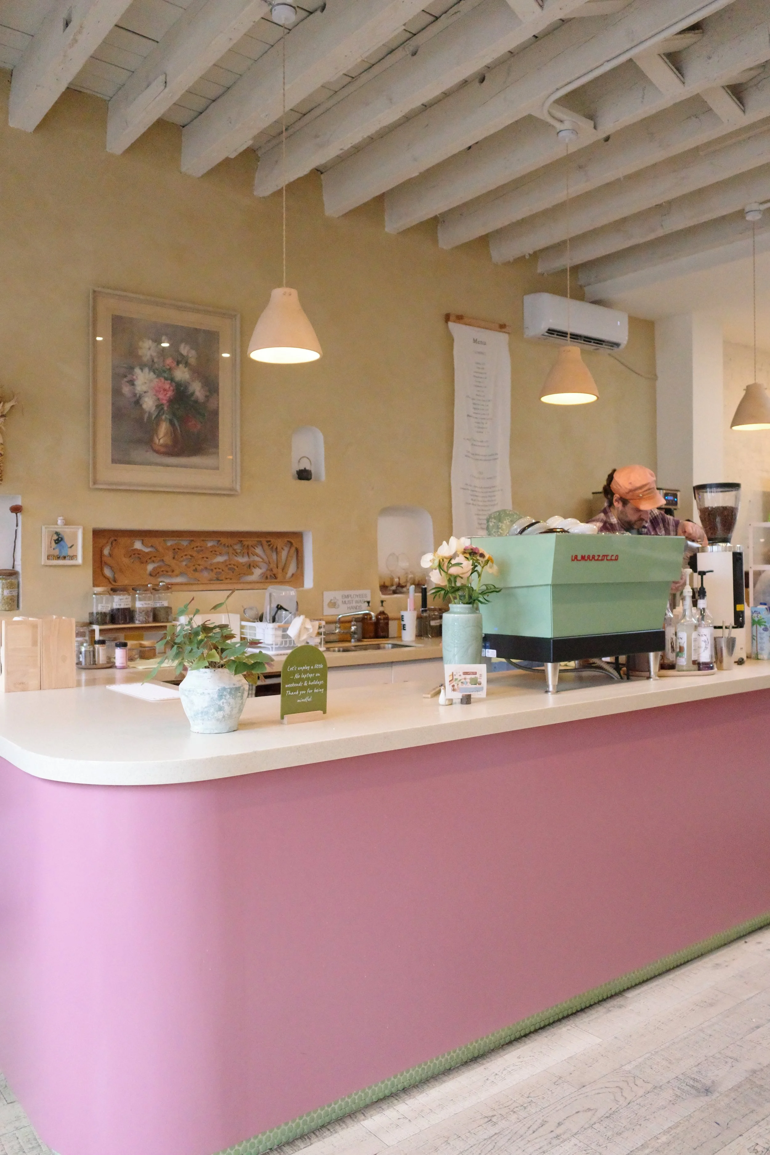 A cafe counter with a pink and green color scheme, decorated with a small potted plant and a green espresso machine. The background shows beige walls, picture frames, a curtain with menu items, and a barista preparing drinks.