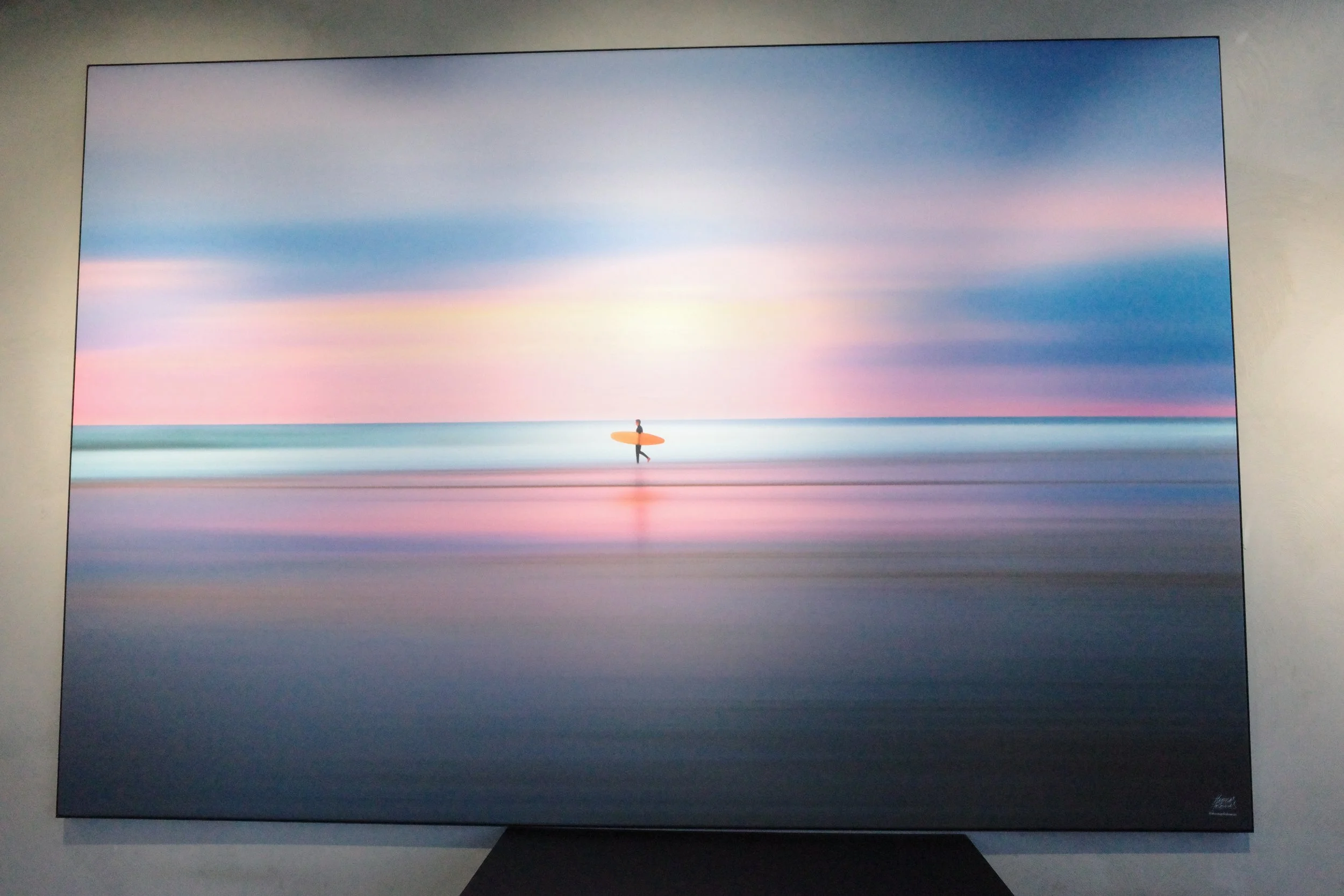 A person holding a surfboard standing on the beach during sunset with a pastel-colored sky.