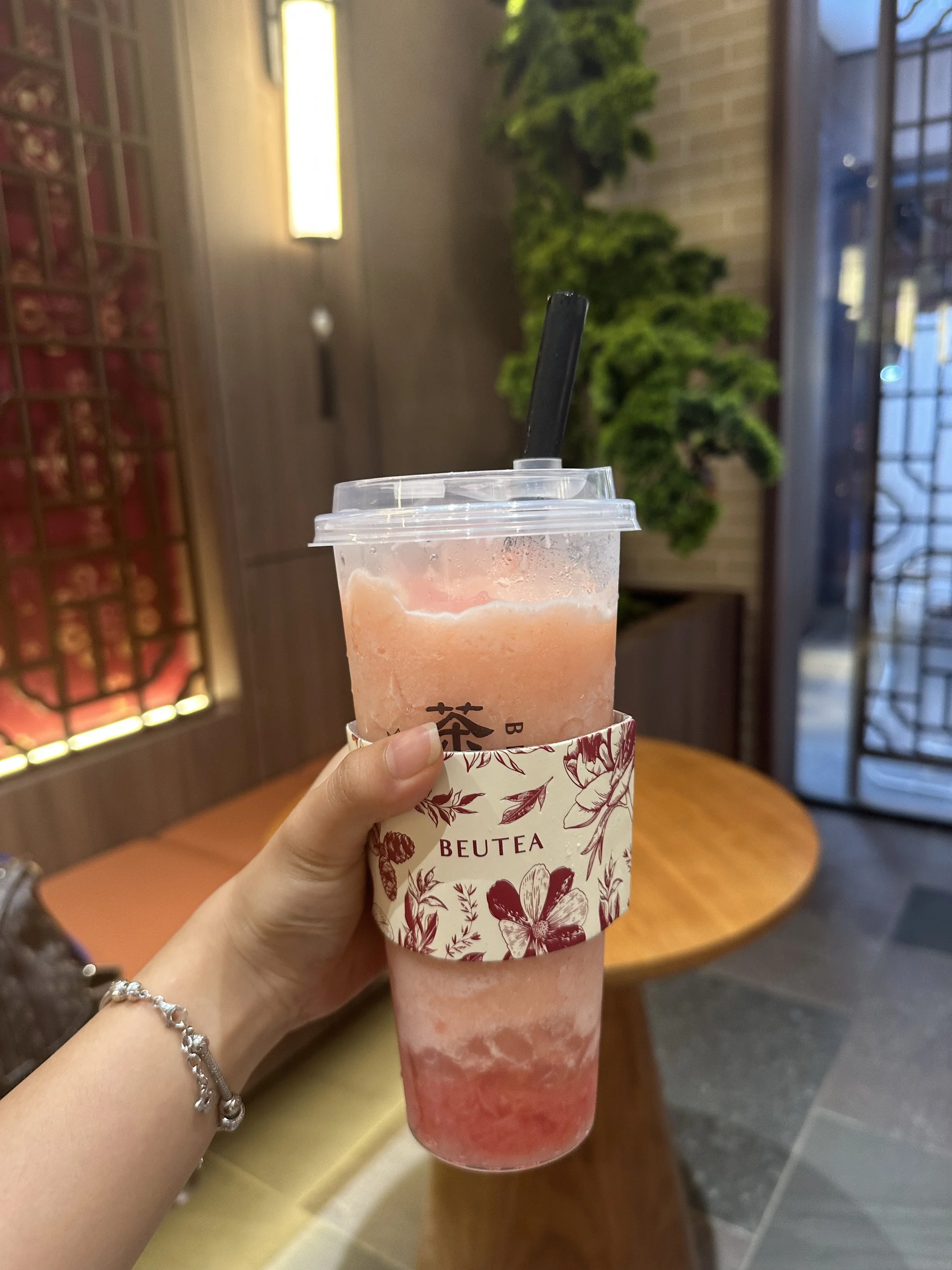 Close-up of a hand holding a pink and red beverage in a plastic cup with a black straw, inside a cafe or restaurant with wooden furniture and greenery in the background.