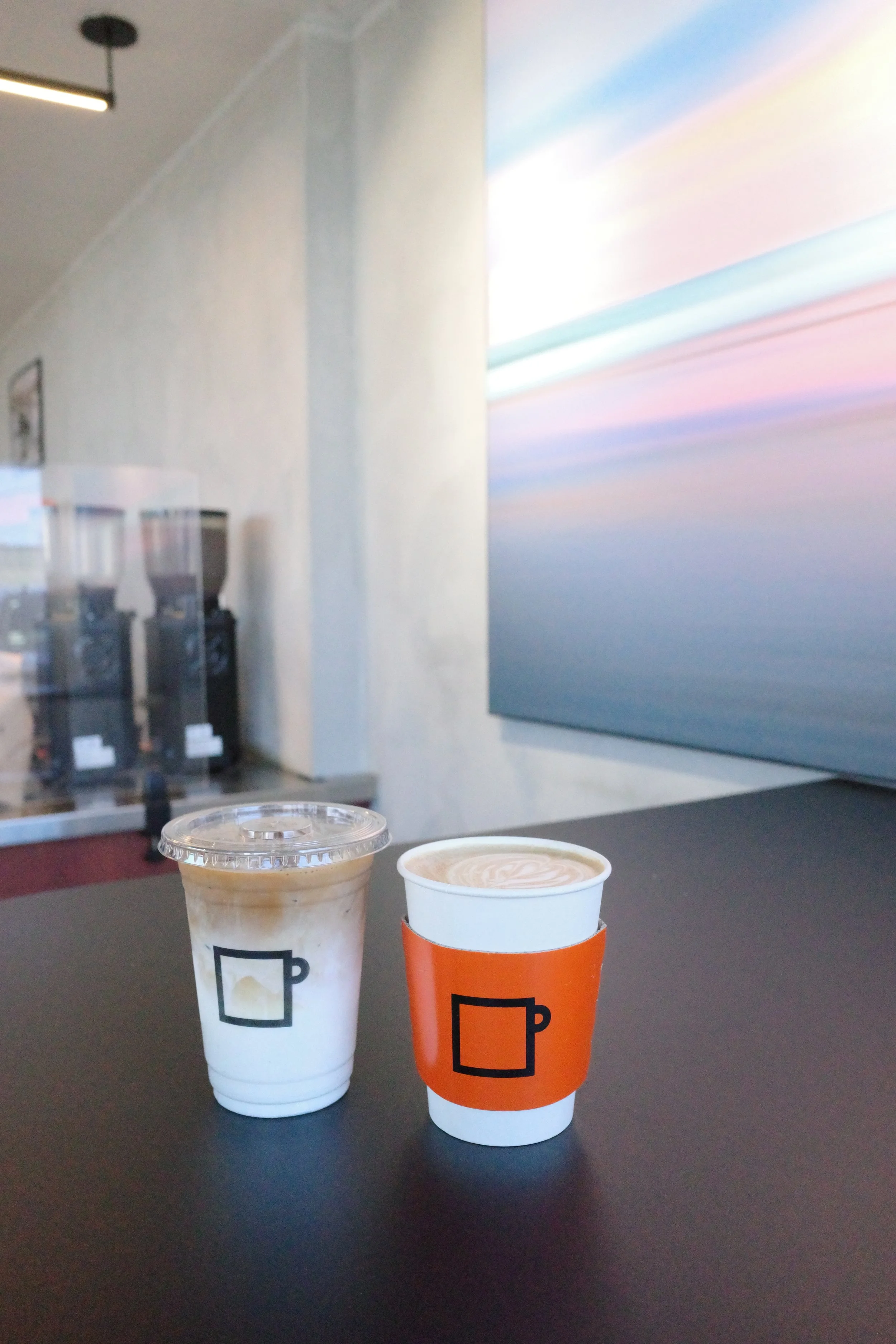 Two coffee cups on a black table, one with a latte and the other with a frappuccino, against a blurry background of an interior space with a wall art piece.