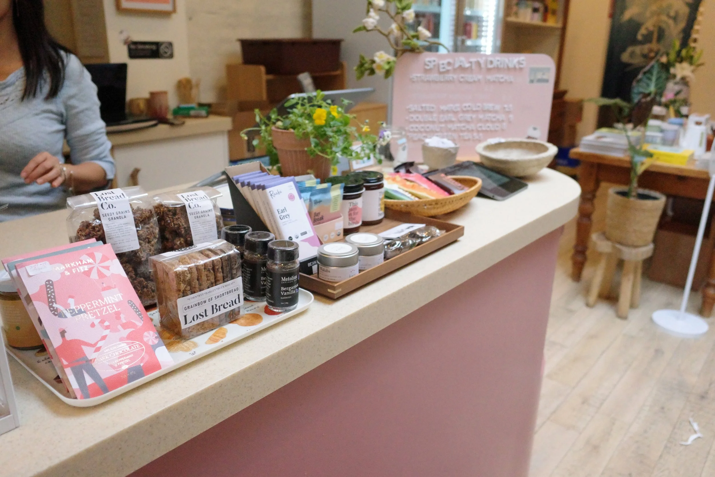 Display of baked goods, snacks, and beverages on a white counter indoors, with a pink sign in the background promoting specialty drinks and a potted plant nearby.