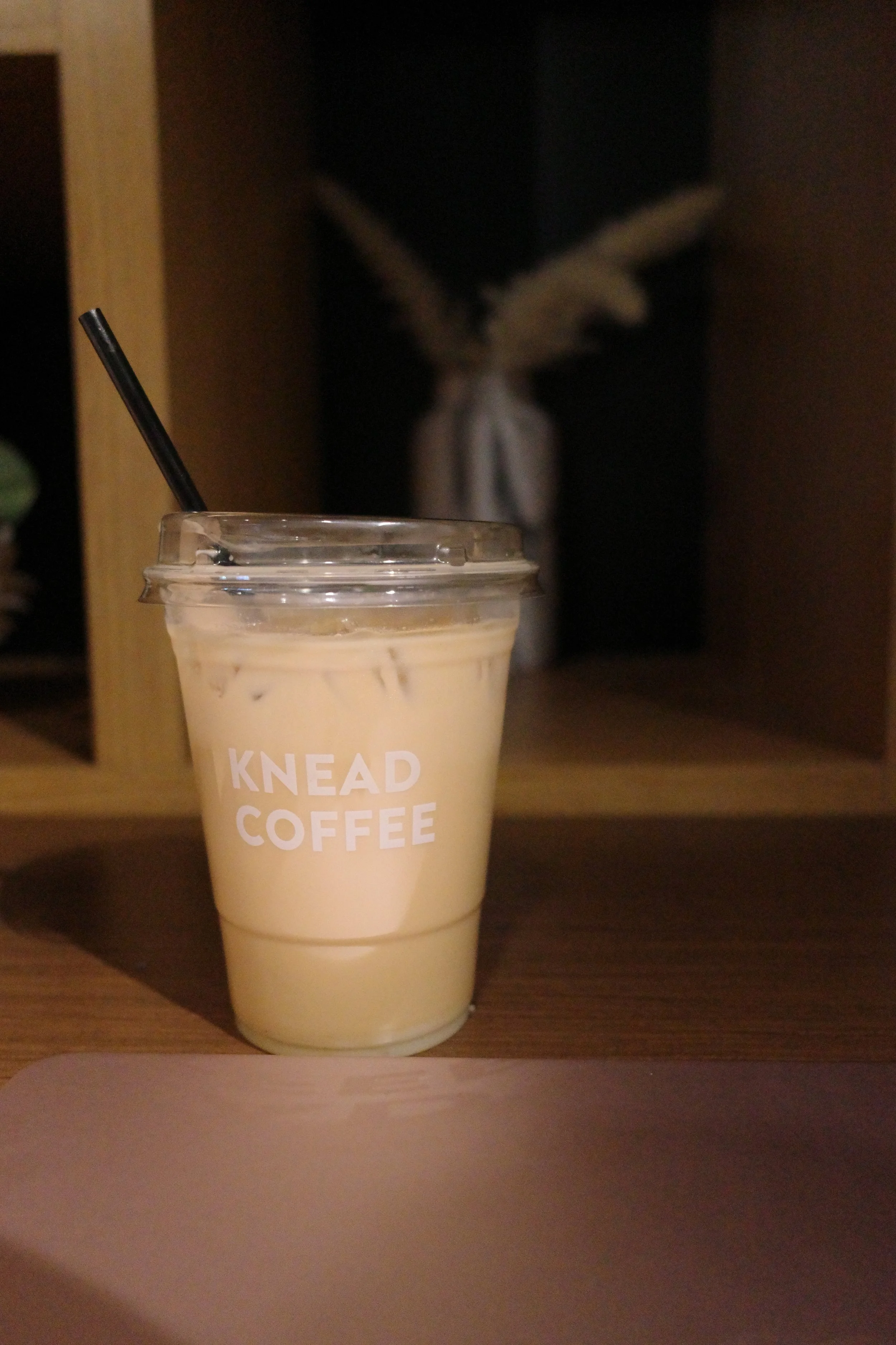 A cold beverage in a clear plastic cup with a black straw, resting on a wooden surface, with a blurred decorative object with white wings in the background.