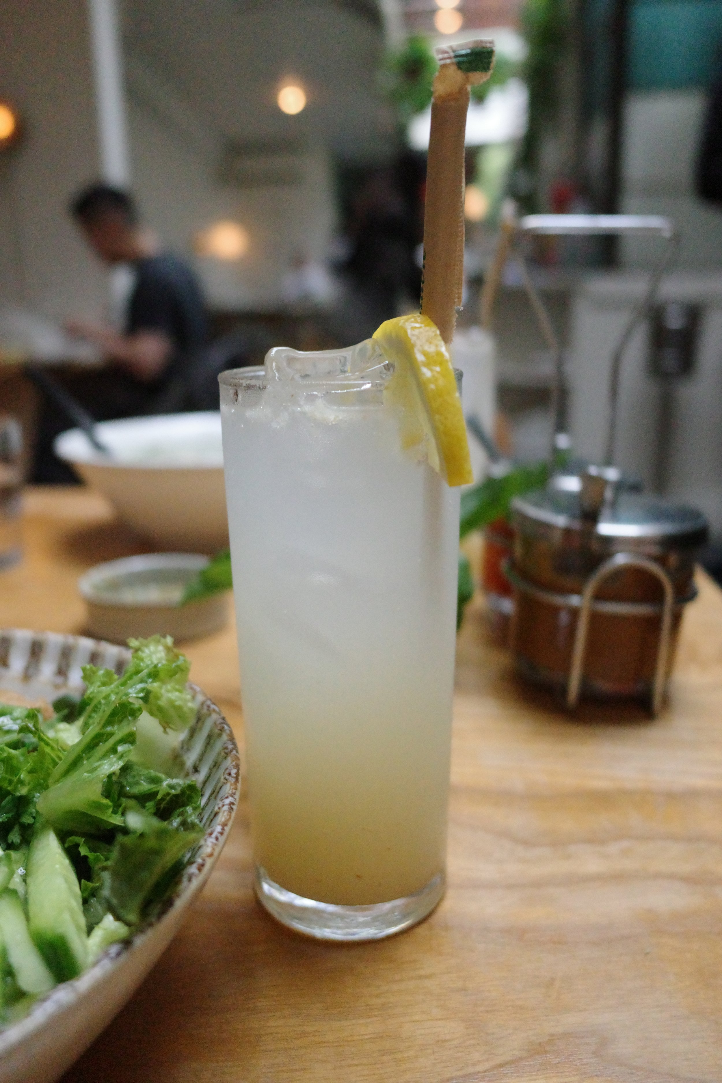 A tall glass of a pale yellow beverage with a lemon slice and a peppermint stick garnishment on the rim, and a bowl of green salad on a wooden table.