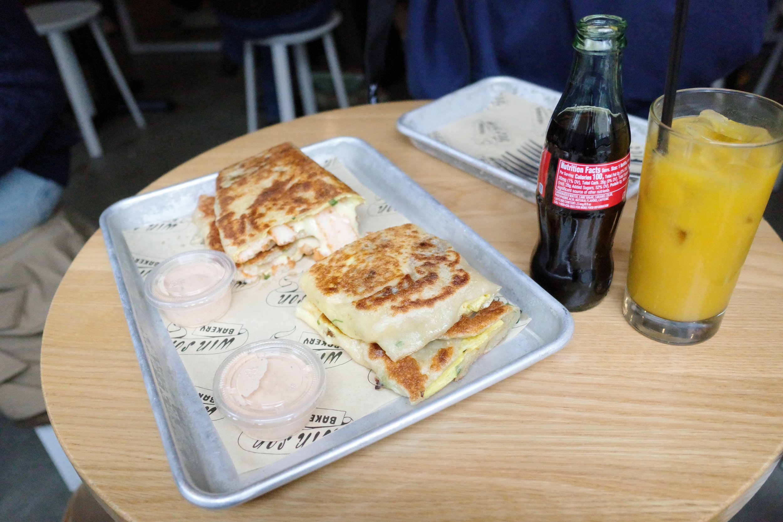 A tray with two pieces of grilled cheese quesadillas, small containers of sauce, a bottle of coke, and a glass of orange juice on a wooden table in a restaurant.
