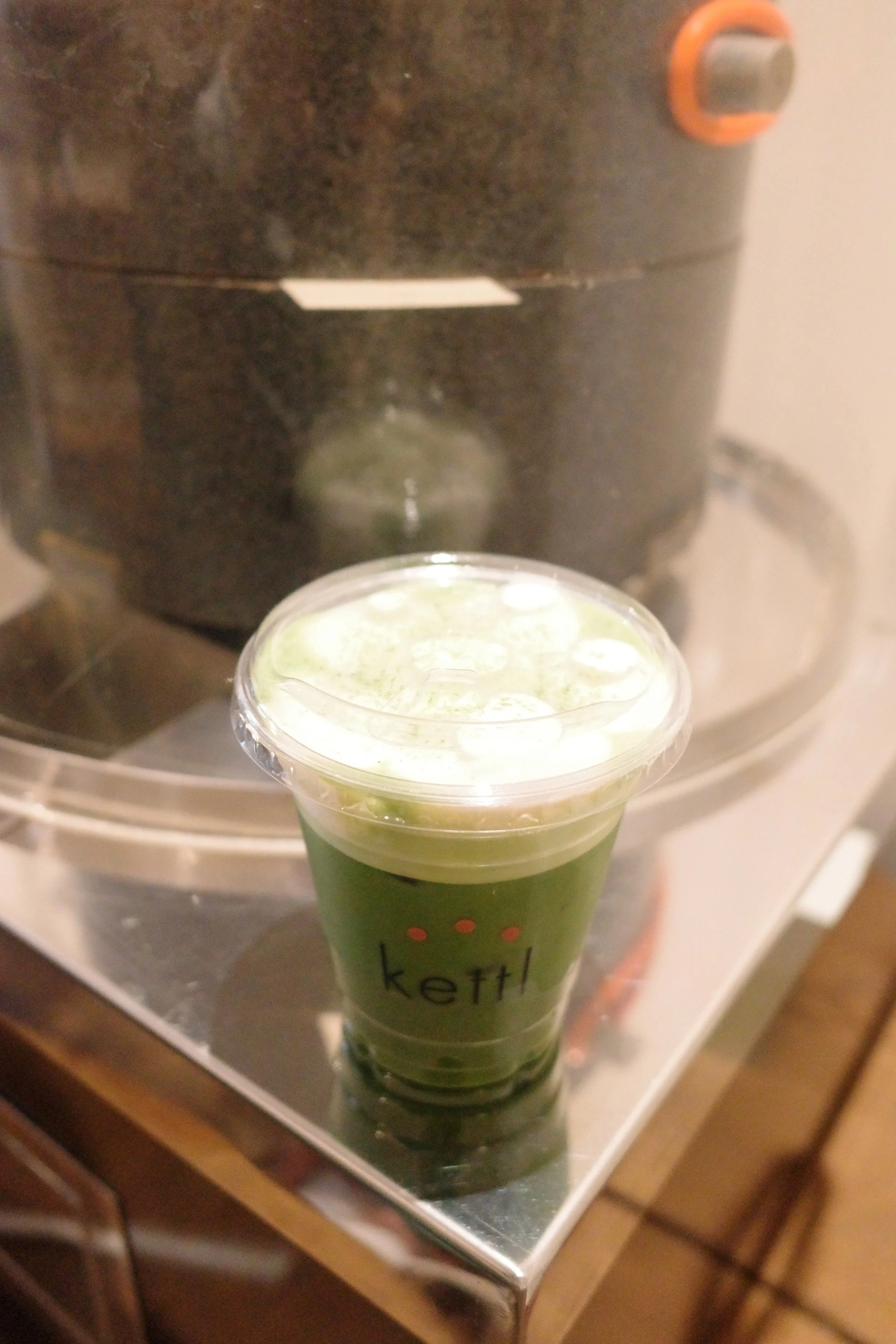 A green iced beverage in a clear plastic cup with a plastic lid, placed on a metal surface with a coffee maker in the background.