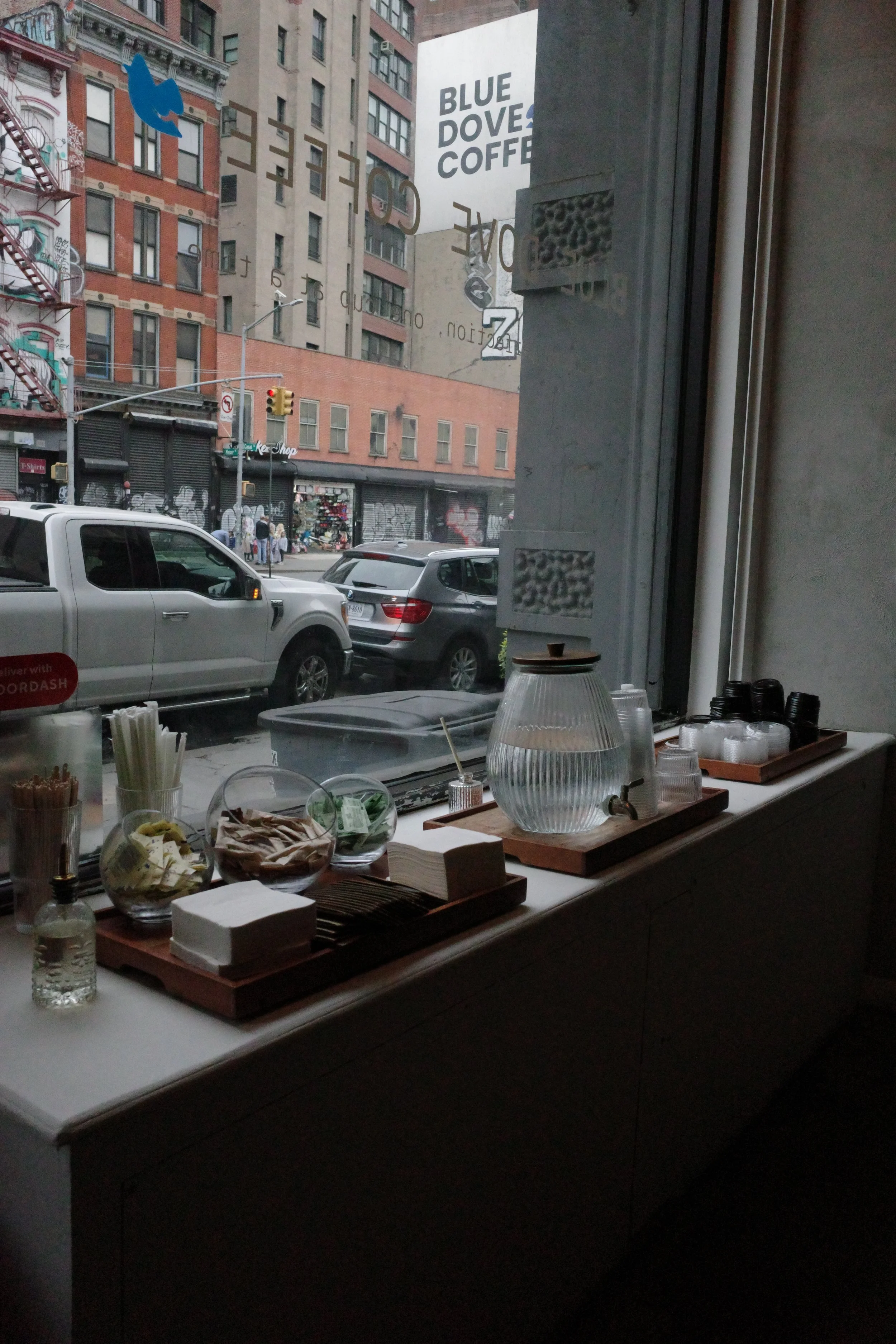 Inside a cafe looking out the window at a city street with parked cars and pedestrians. Exterior buildings have graffiti, signs, and a large sign for Blue Doves Coffee.