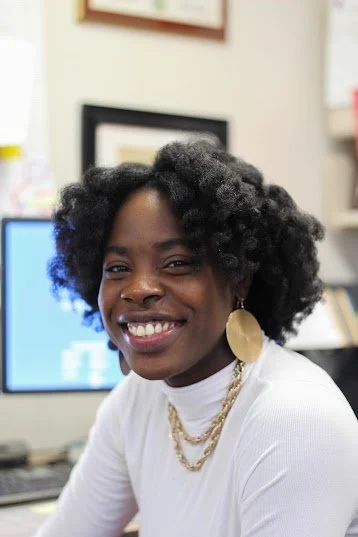 A woman with natural curly hair smiling in an office setting, wearing a white top, large gold earrings, and layered gold necklaces.