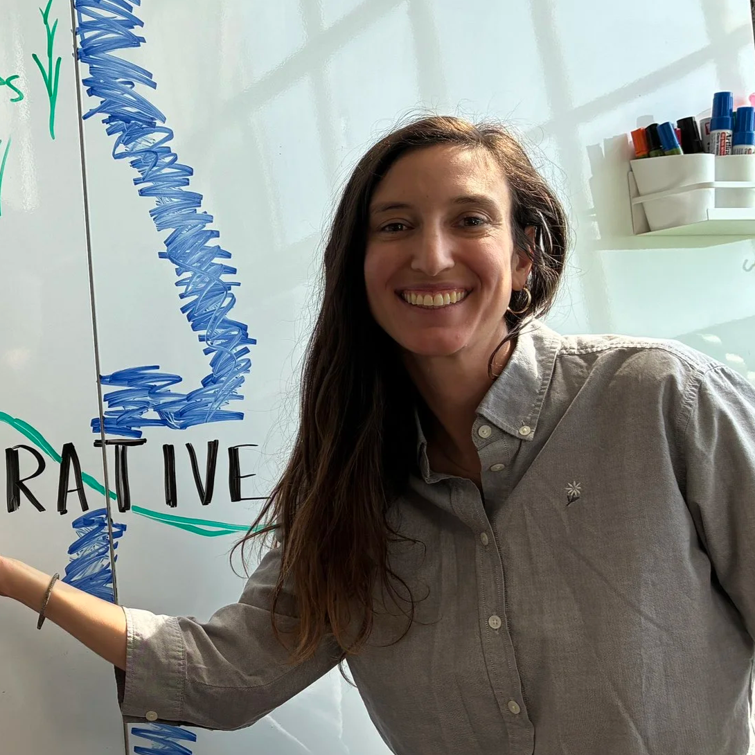 A woman smiling and pointing at a whiteboard with a graph and the word 'CREATIVE' written on it.