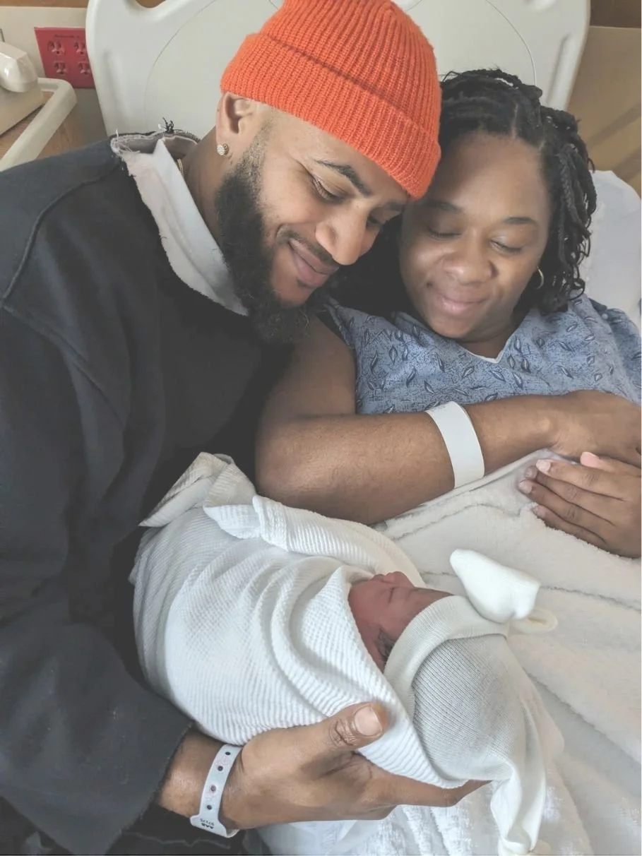 Newborn baby wrapped in a white blanket being held by a man wearing a black jacket and red beanie, with a woman in a hospital gown and wristband lying in a hospital bed beside them.
