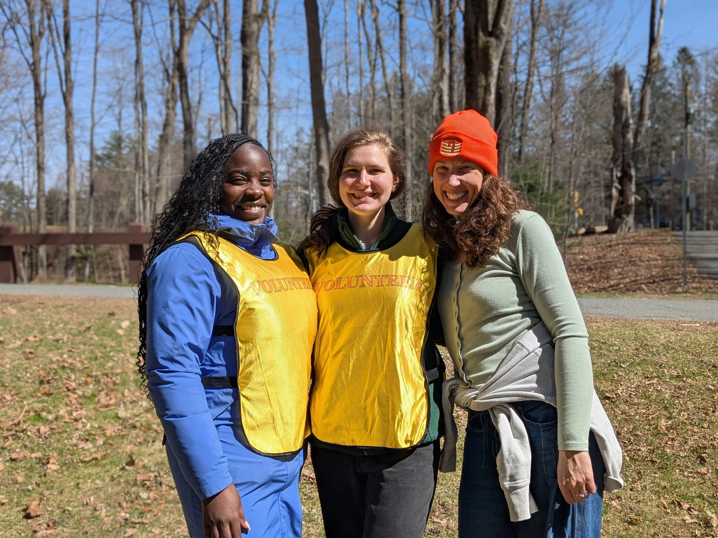 Three women standing outdoors in a park, smiling at the camera. Two women are wearing yellow volunteer vests, one of whom is wearing a blue jacket. The third woman is wearing a light green top and an orange beanie. The background shows leafless trees and a clear blue sky.