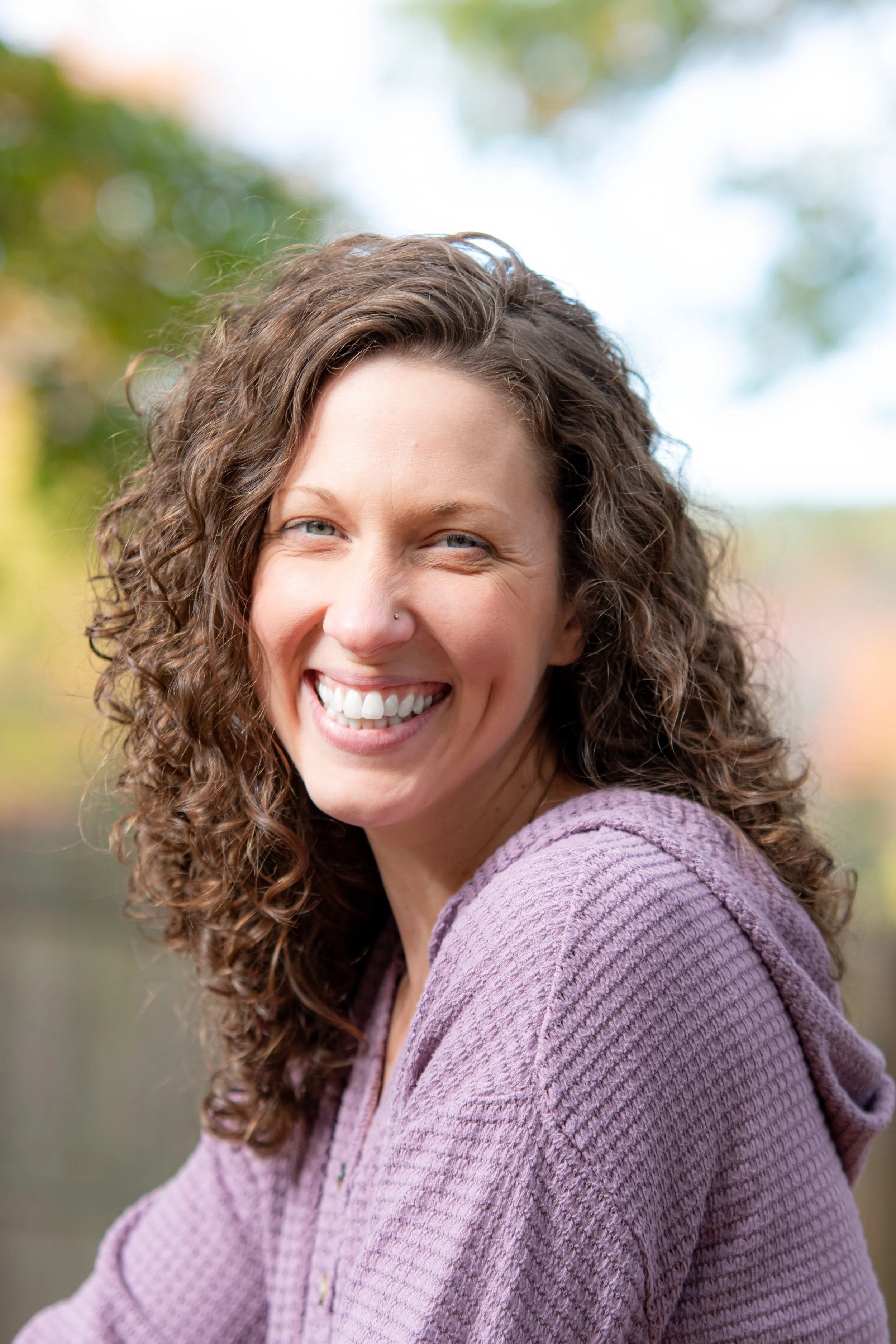 A smiling woman with curly brown hair and light skin, wearing a purple sweater, outdoors with blurred green foliage in the background.