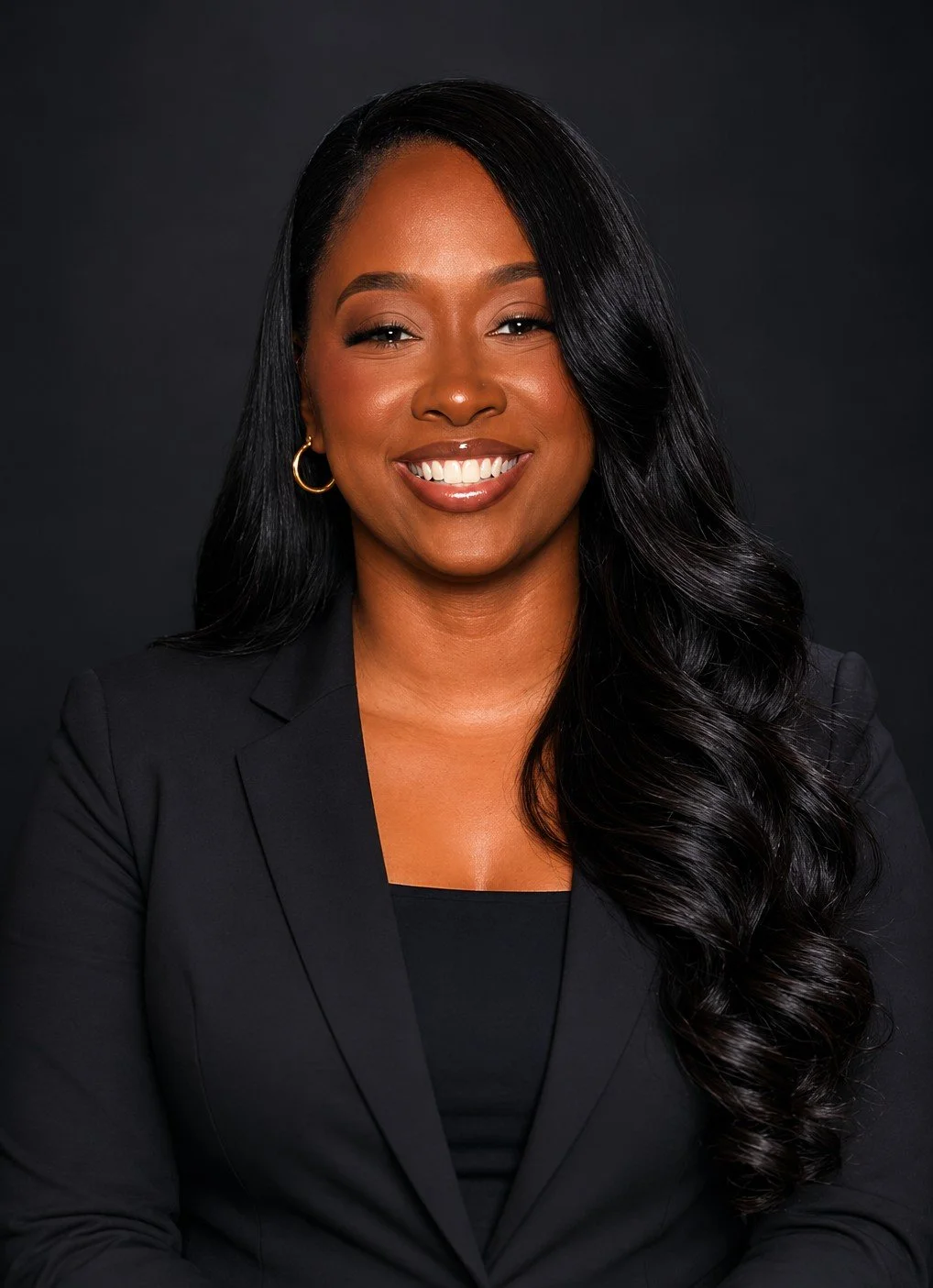A smiling woman with long black hair styled in loose curls, wearing a black blazer and gold hoop earrings, against a dark background.