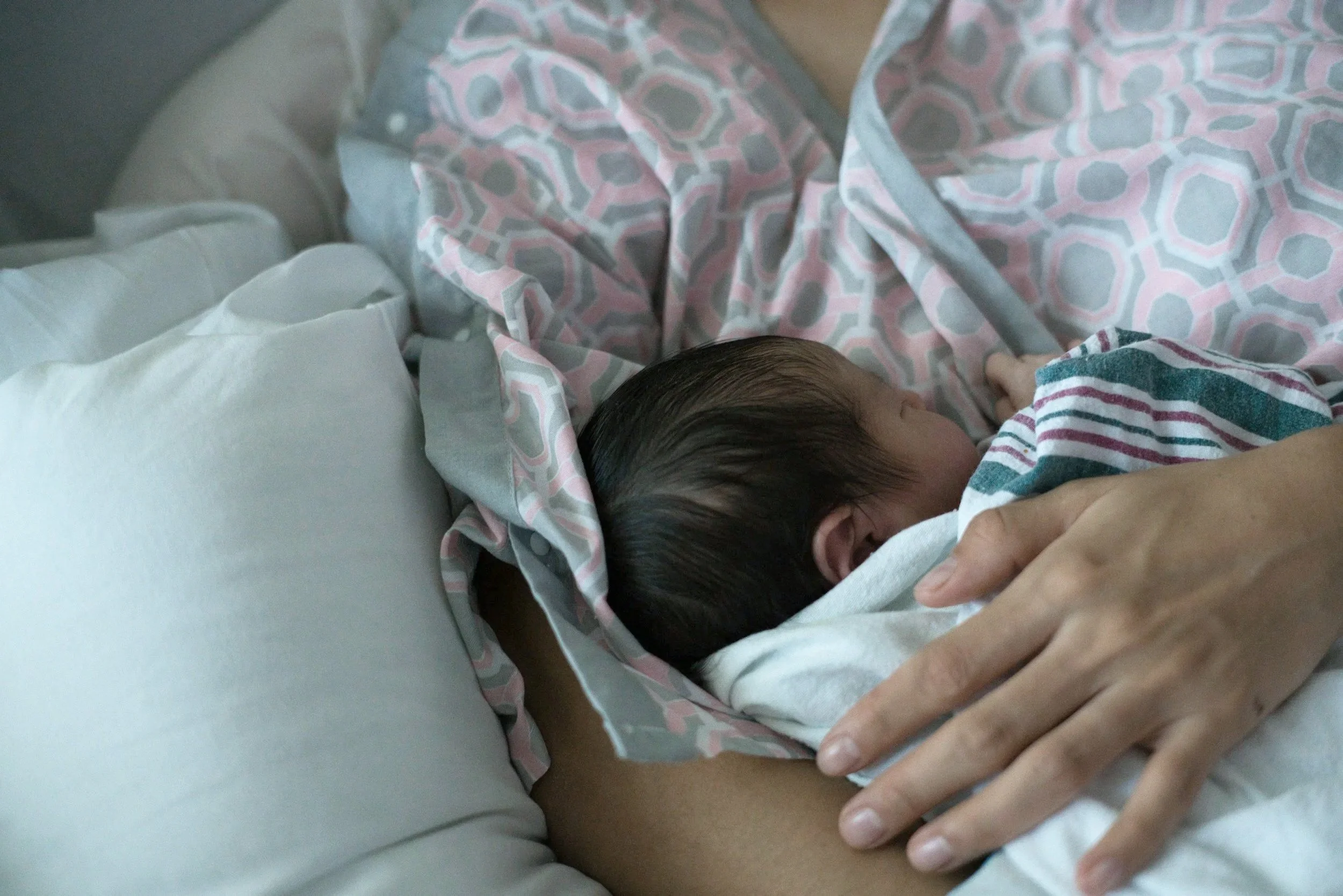 A newborn baby with dark hair sleeping on a blanket, with adult hands gently holding the baby's shoulder.