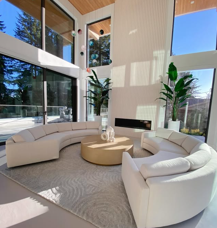 Modern living room with large floor-to-ceiling windows, two white curved sofas, a round beige coffee table with decorative objects, and tall green plants in white pots, with sunlight casting shadows inside.