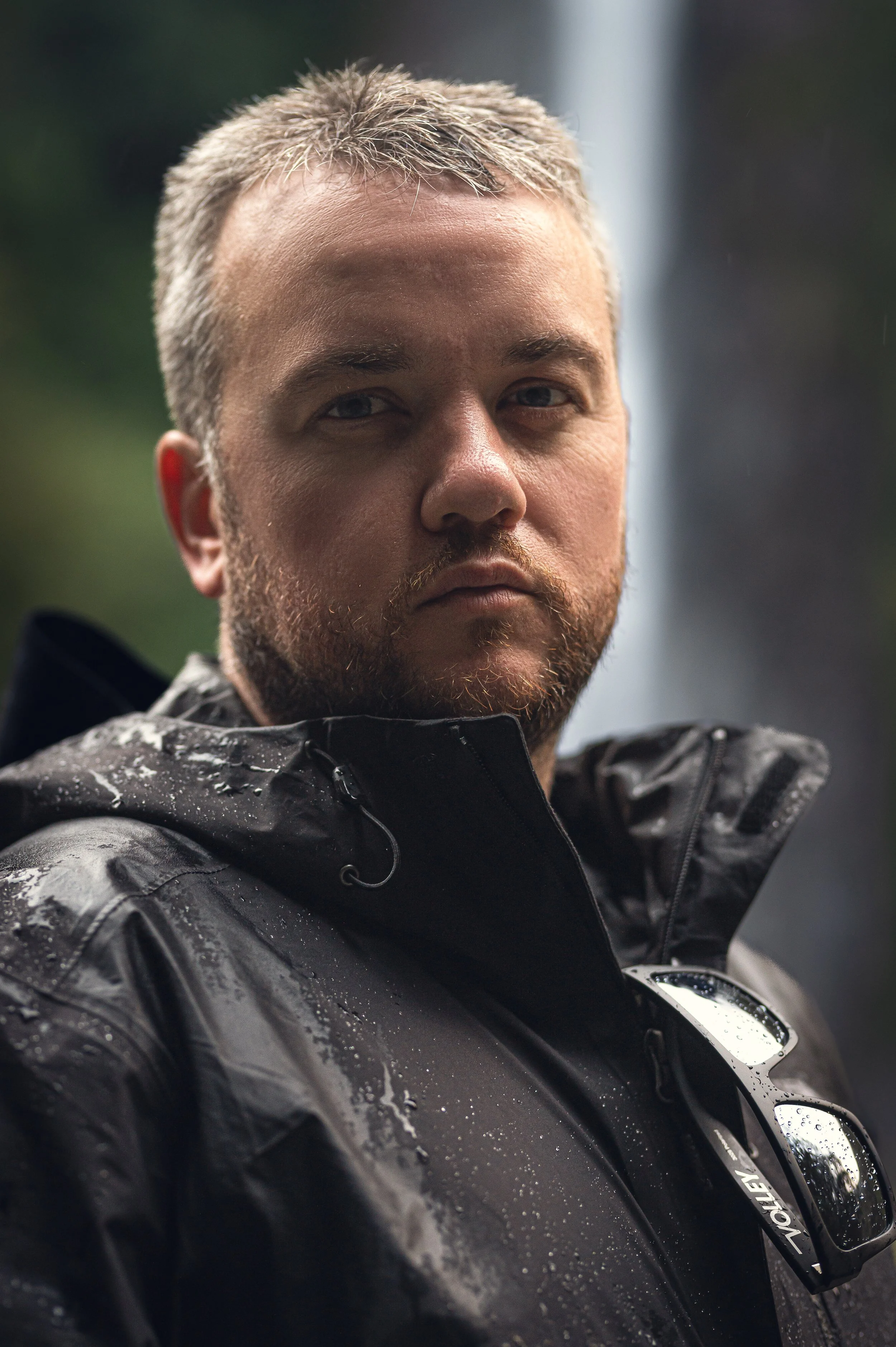 Close-up of a man with short, light-colored hair and a beard, wearing a black waterproof jacket with water droplets, standing outdoors in a rainy environment.