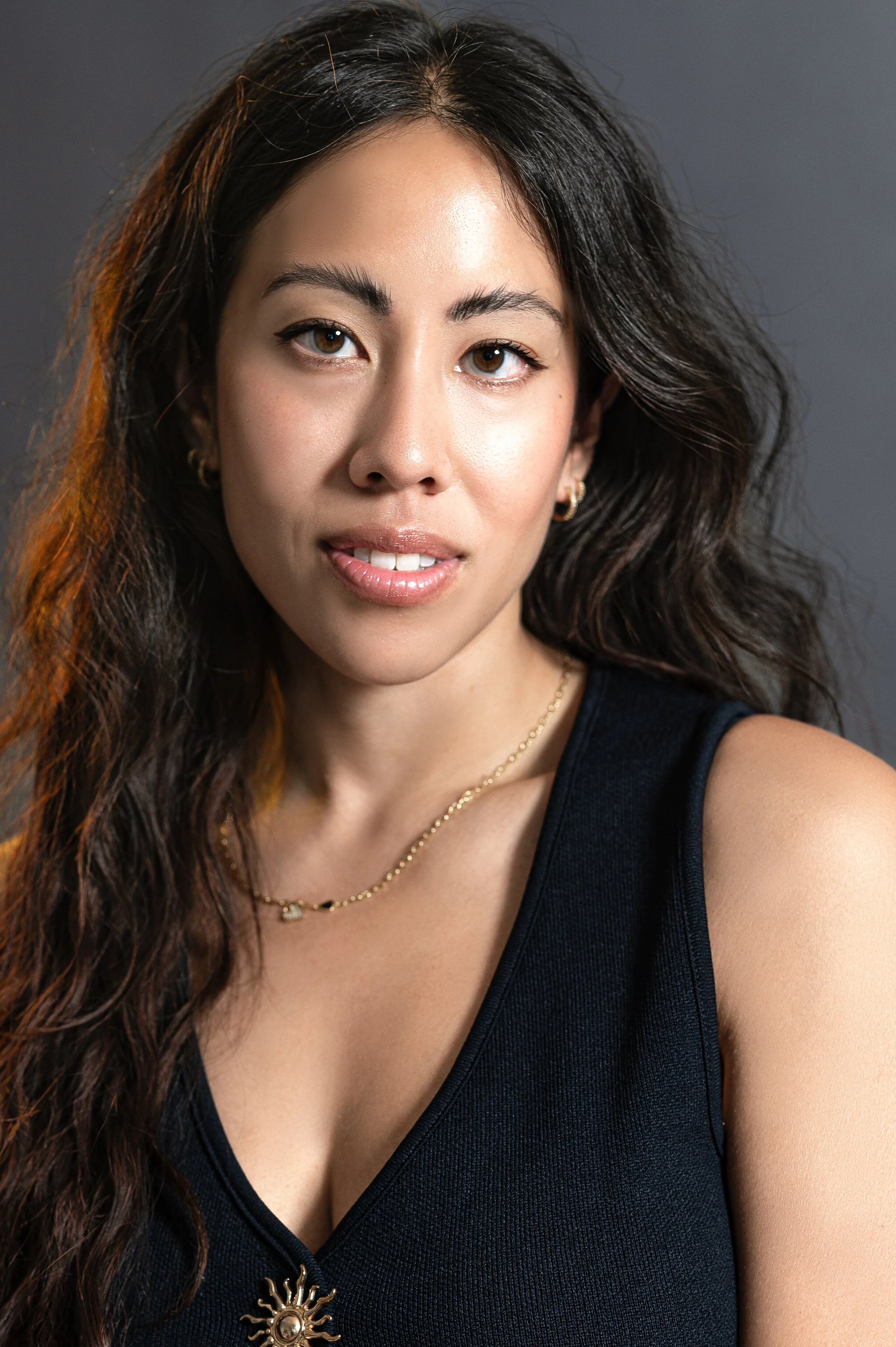 Close-up of a woman with long, wavy brown hair wearing a black sleeveless top, gold jewelry, and subtle makeup against a gray background.