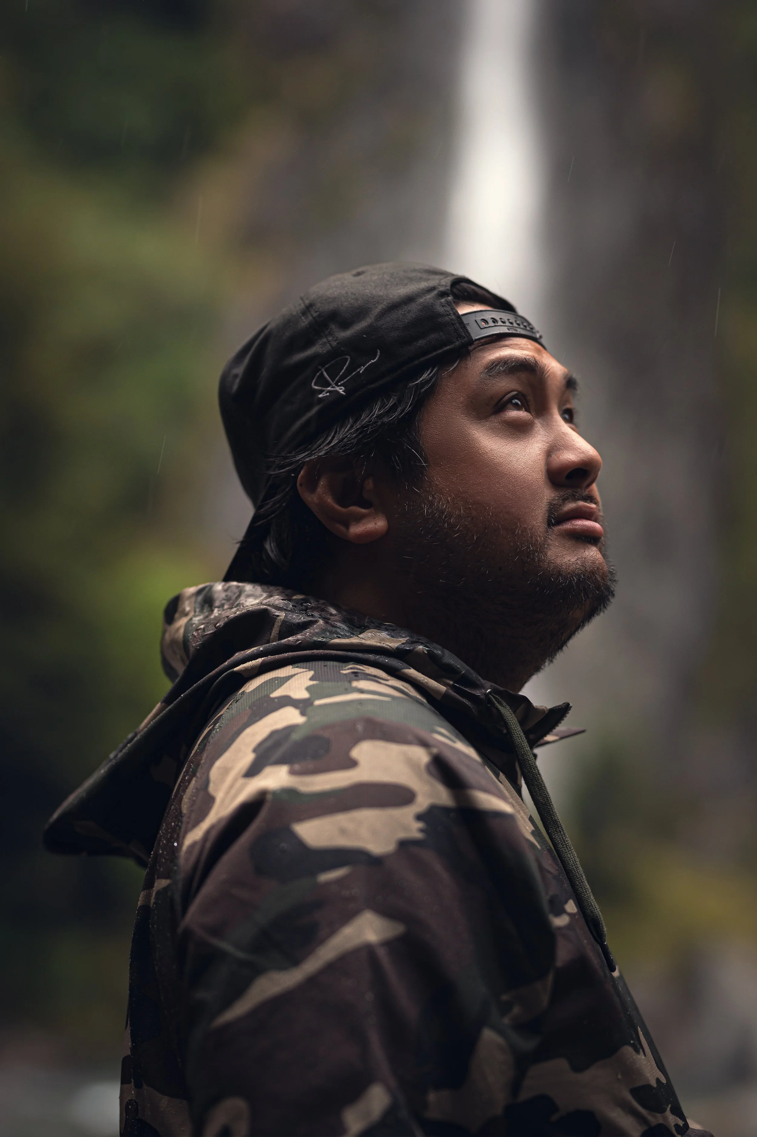 A man with a beard and long dark hair, wearing a black cap backwards and camouflage jacket, looking upward with a waterfall in the background during rainy weather.