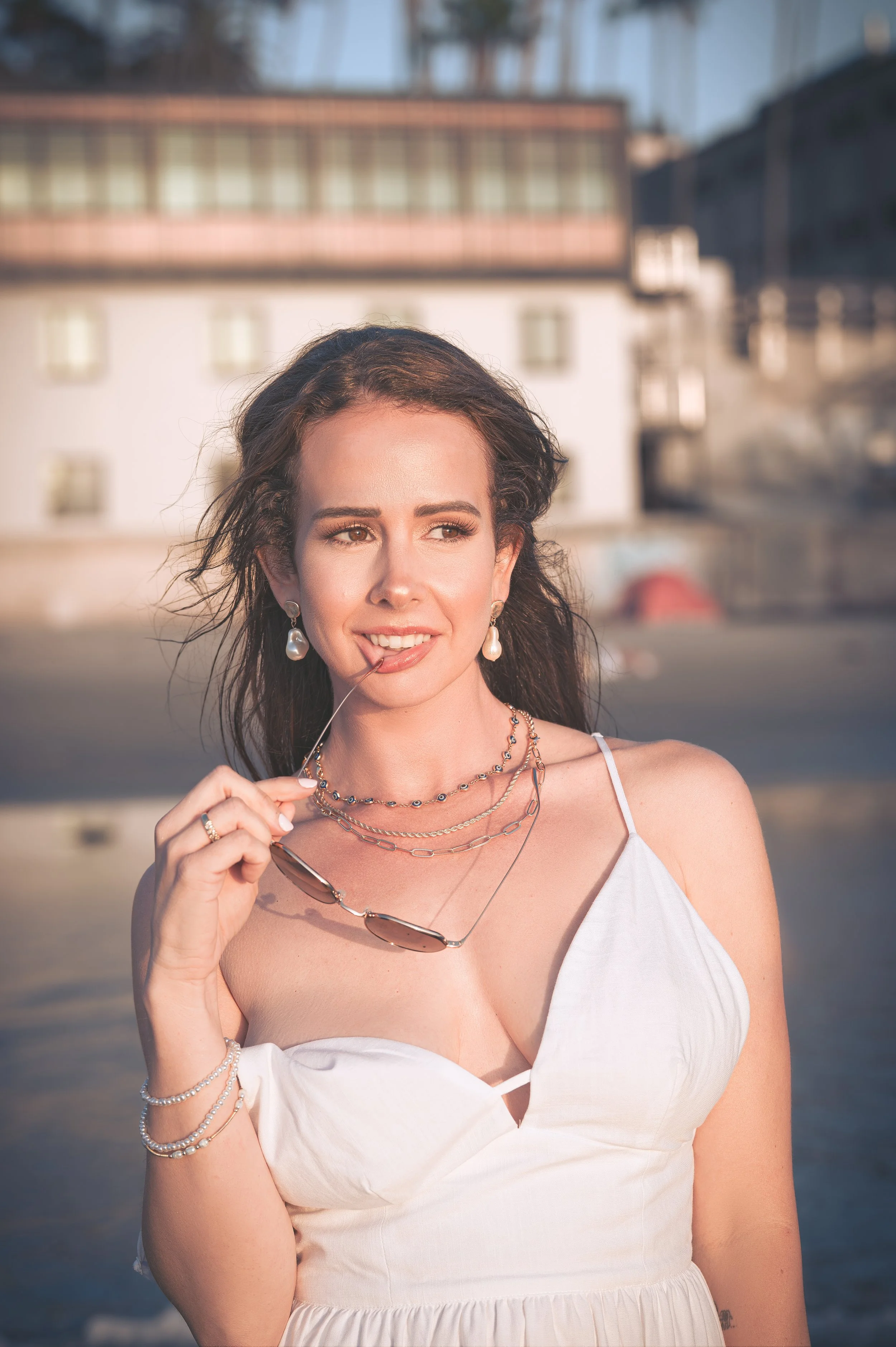 A woman with brown hair wearing a white dress, pearl earrings, and layered necklaces, holding sunglasses near her face, standing outdoors in sunlight with blurred buildings in the background.