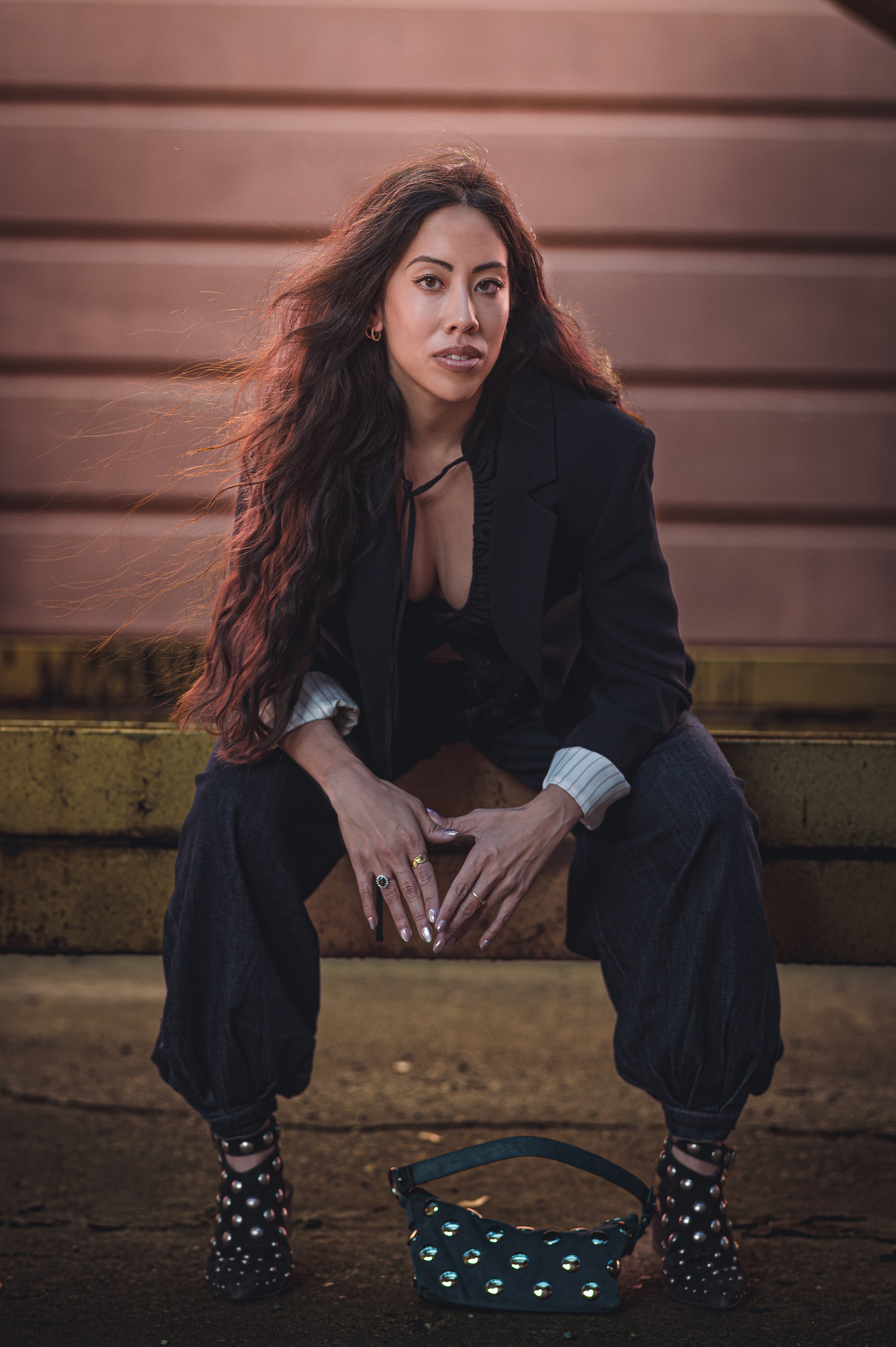 A woman with long, wavy dark hair sitting on a wooden bench outdoors, wearing a black blazer, dark pants, and studded black ankle boots. She has jewelry on her fingers and earrings. There is a matching studded handbag at her feet.