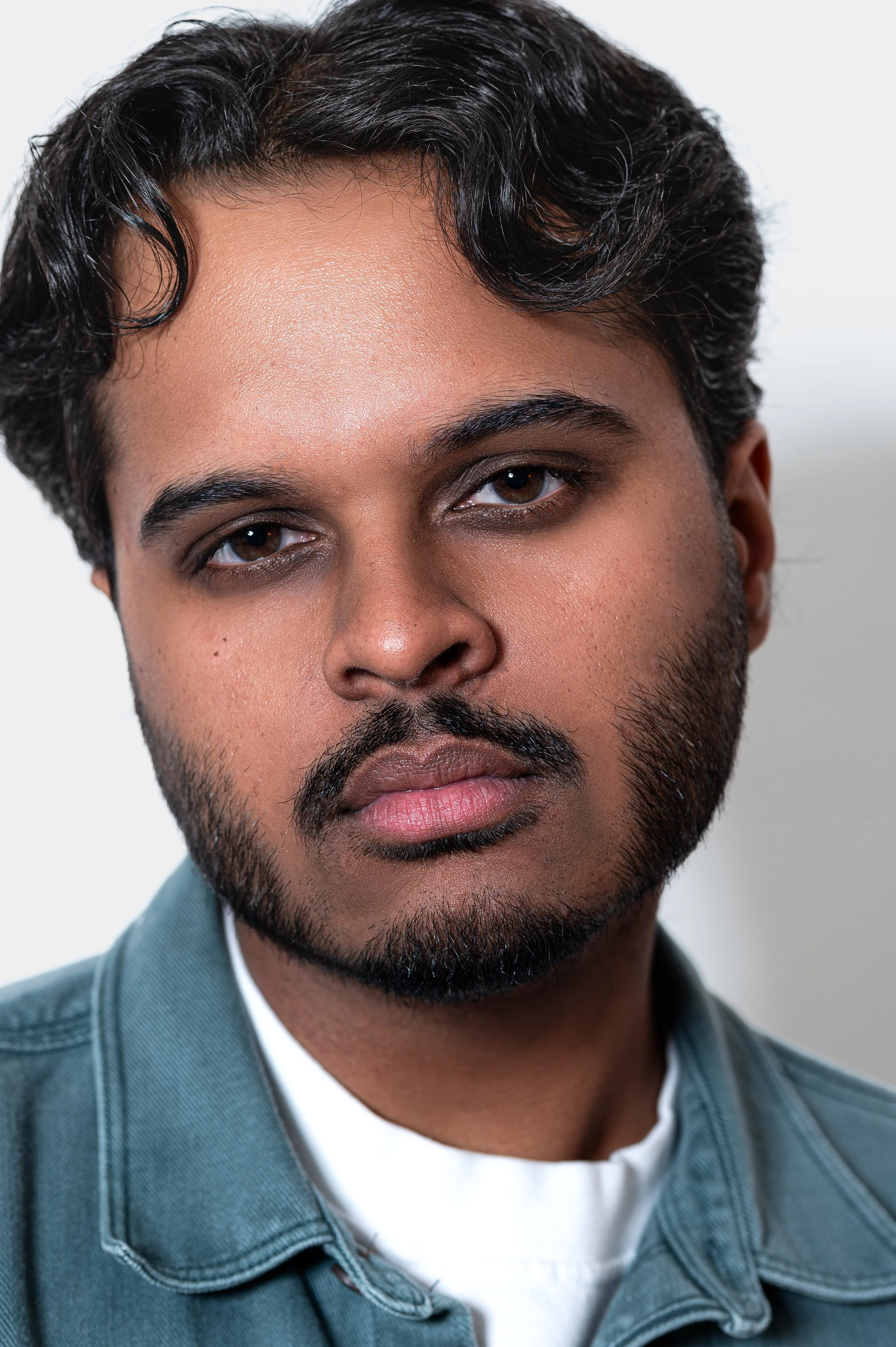 Close-up portrait of a man with dark hair, brown eyes, and a beard, wearing a denim jacket over a white t-shirt, against a plain white background.