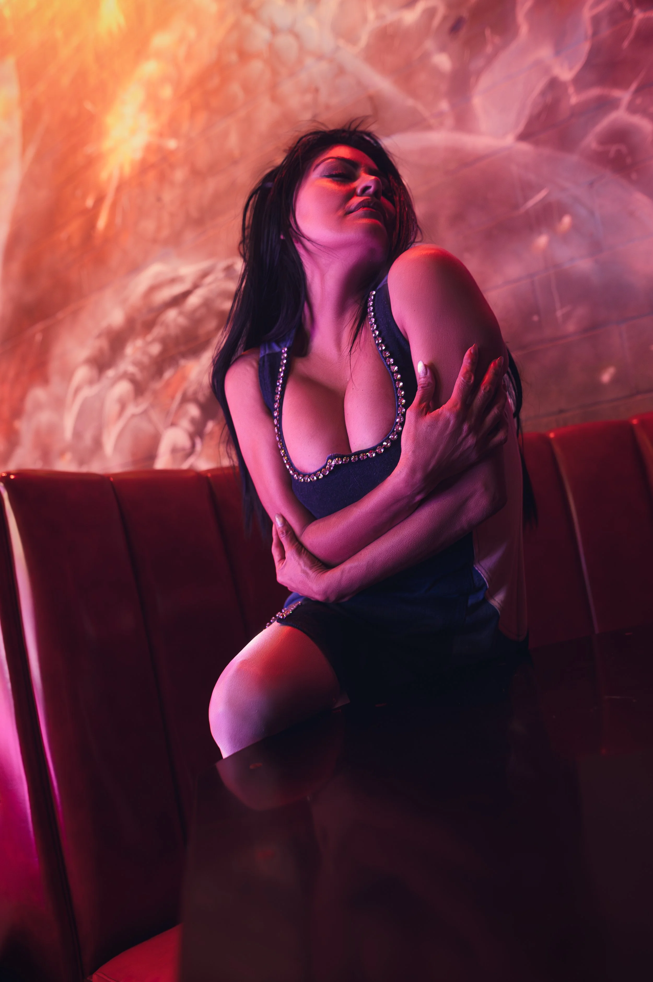 A woman with dark hair and makeup, wearing a black top with embellished trim, sitting on a red booth seat against a marbled wall with pink and purple lighting, her eyes closed and arms crossed over her chest.