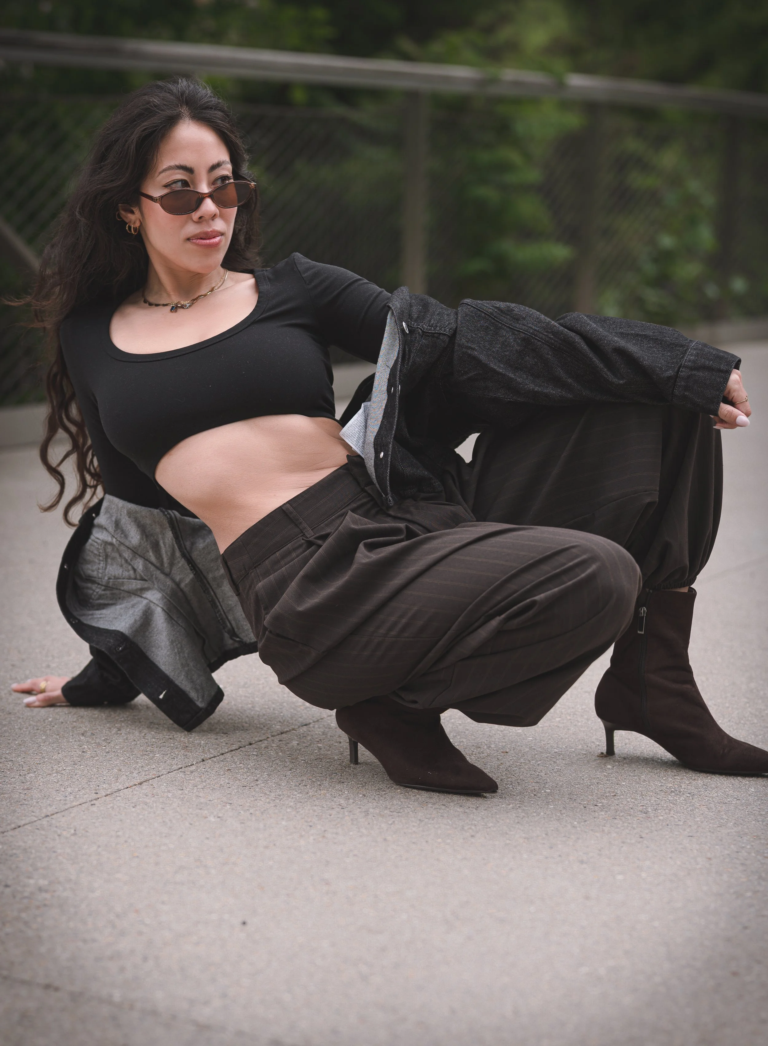 A woman with long dark hair and sunglasses is crouching on the ground, wearing a black top, a denim jacket, brown striped pants, and black heeled boots, outdoors with greenery and a fence in the background.