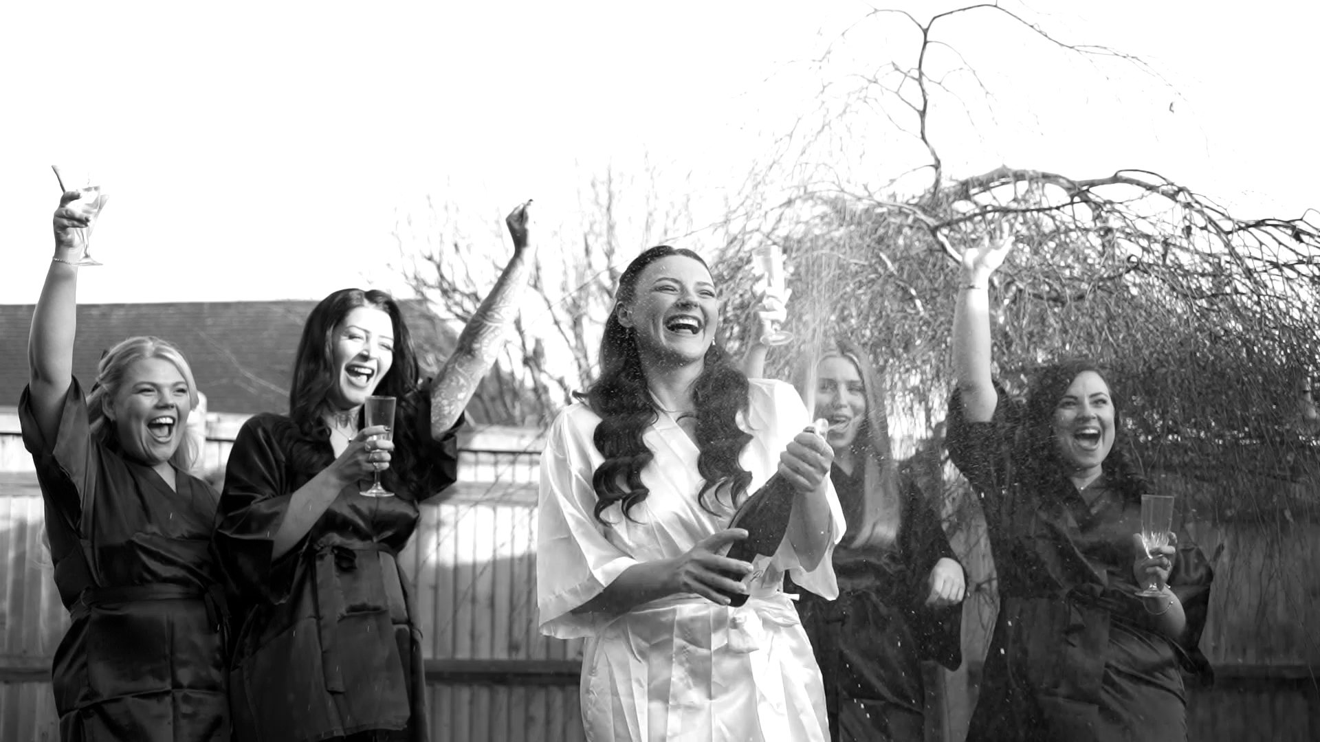 Group of five women celebrating outdoors, smiling and raising glasses, with one woman in the center holding a sparkler.