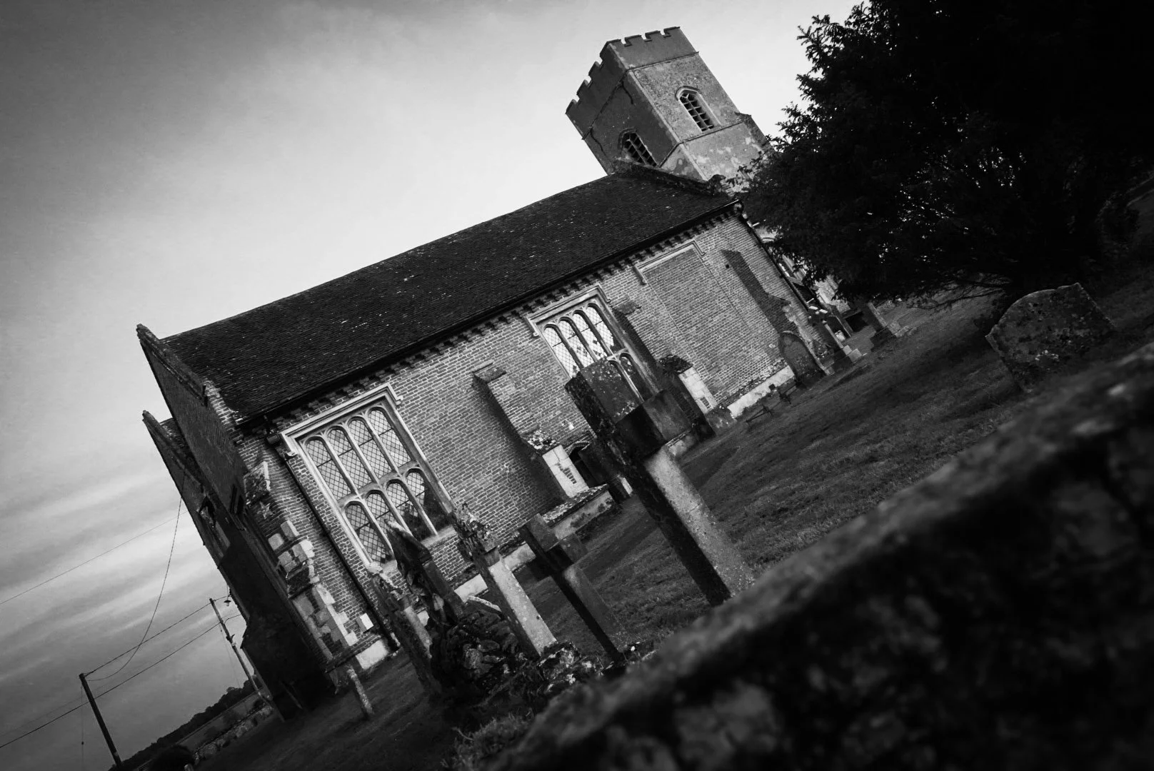 Black and white photo of a church with a tall tower, arched windows, a sloped roof, and surrounding yard with old gravestones and a tree.