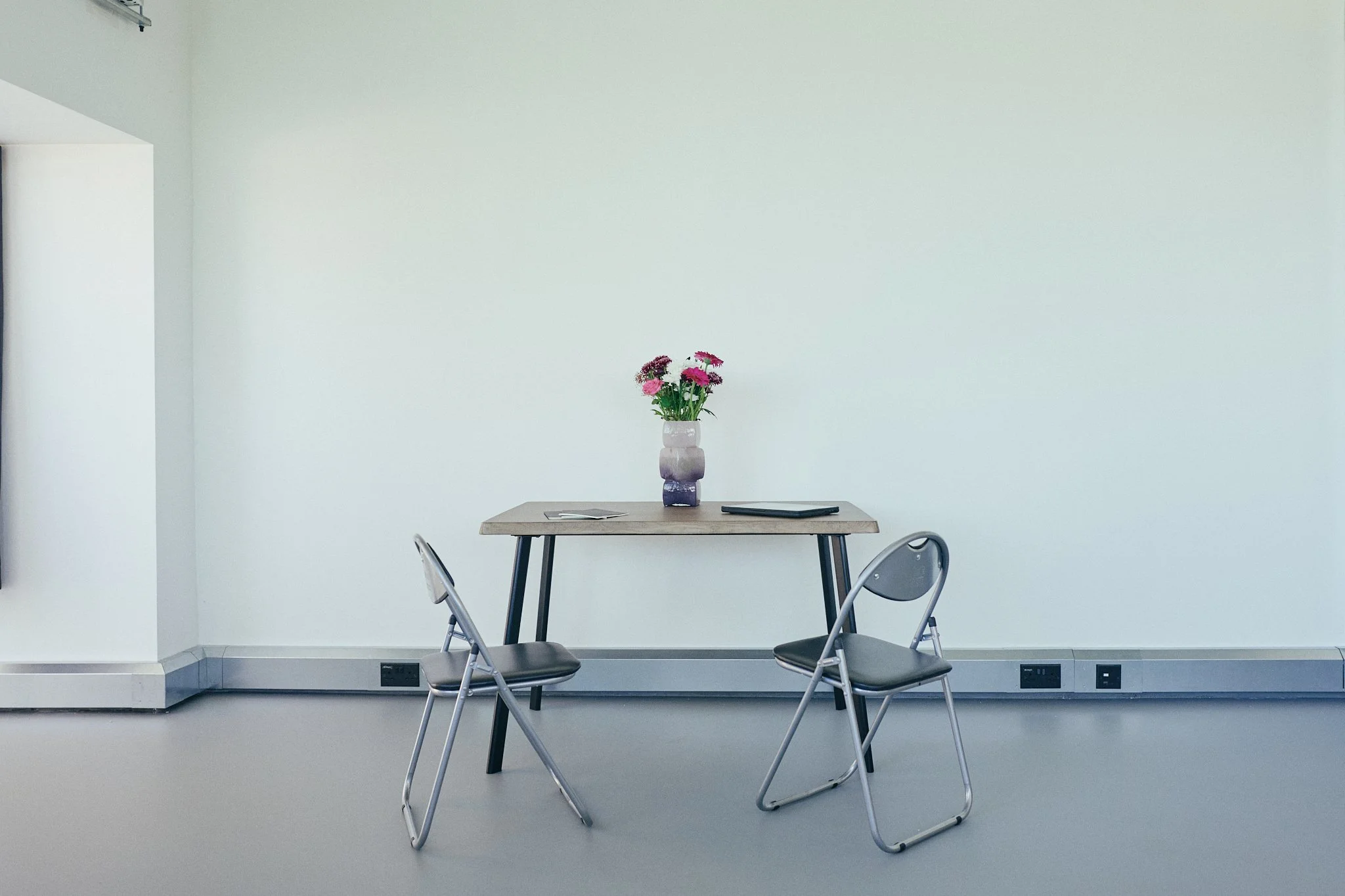 A minimalist room with light gray walls and flooring, featuring a wooden table with black legs. On the table is a pink, purple, and white flower arrangement in a ceramic vase, a black notebook, and a piece of paper. Two metal chairs with black seats 