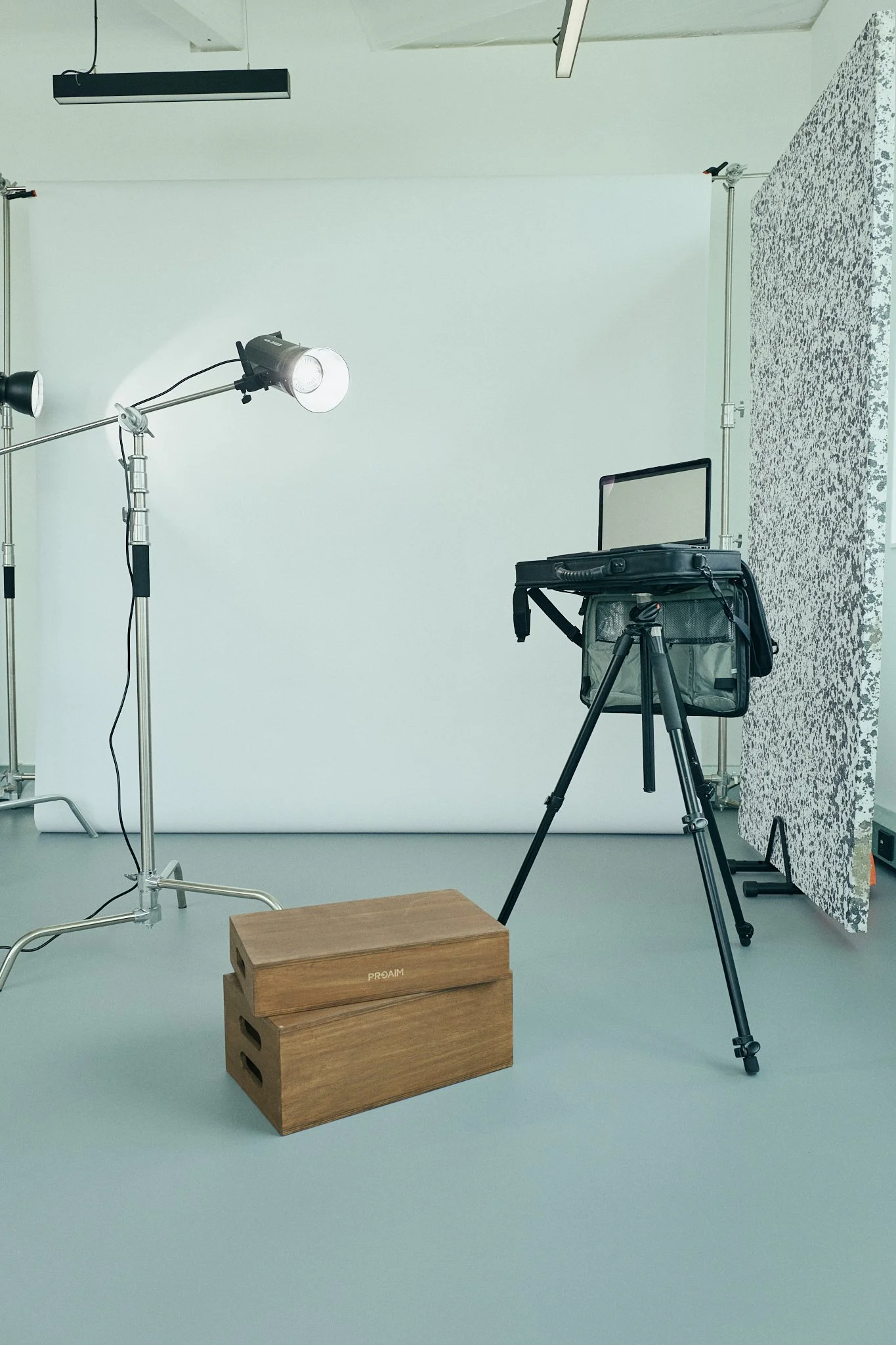 Photo of a photography studio setup with a white backdrop, professional lighting, a camera on tripod, a wooden box, and a patterned wall on the right.