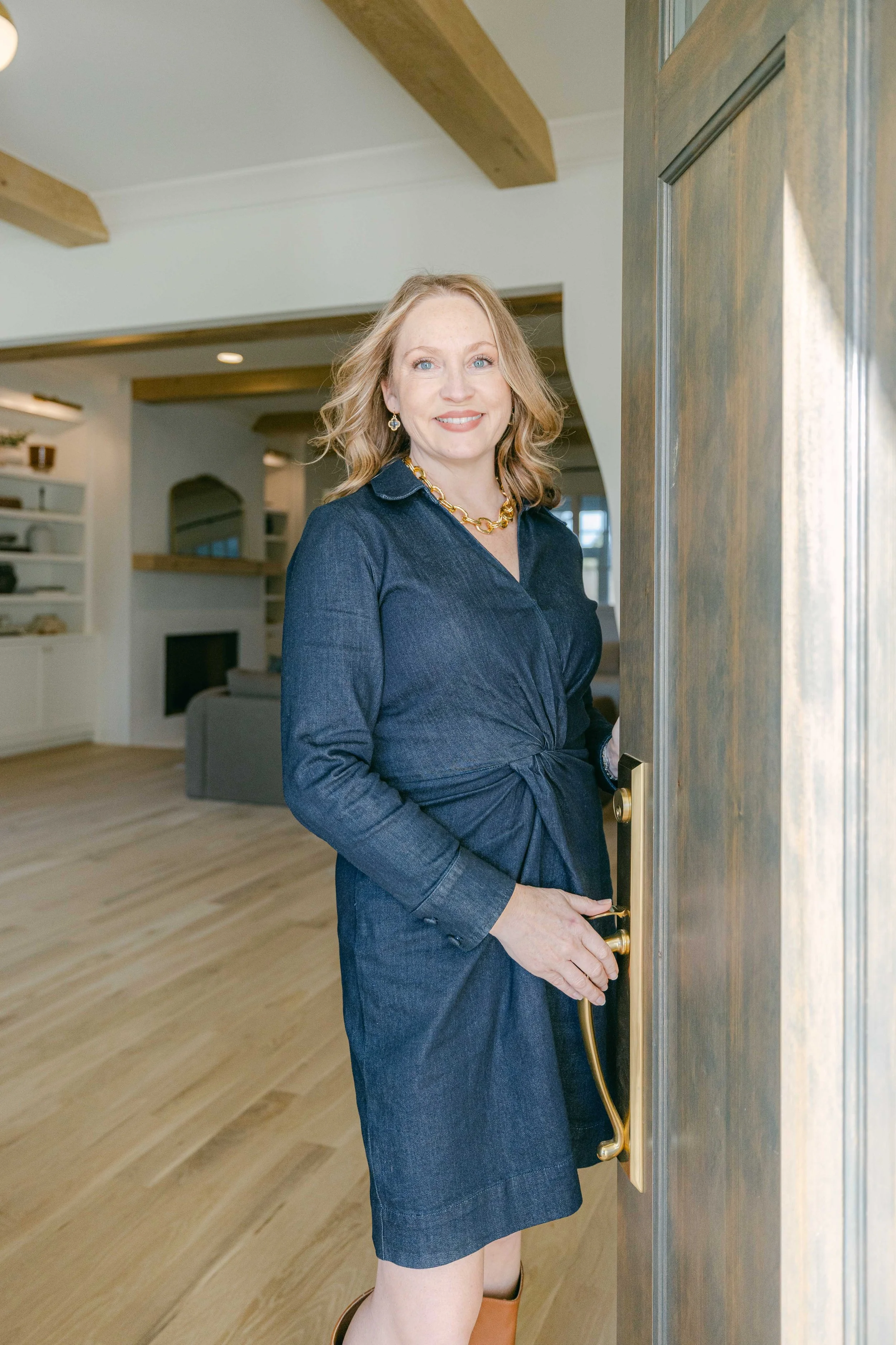 A woman with blonde hair, blue eyes, wearing a dark blue dress, gold jewelry, and brown boots, standing at an open wooden door in a bright, modern living room.