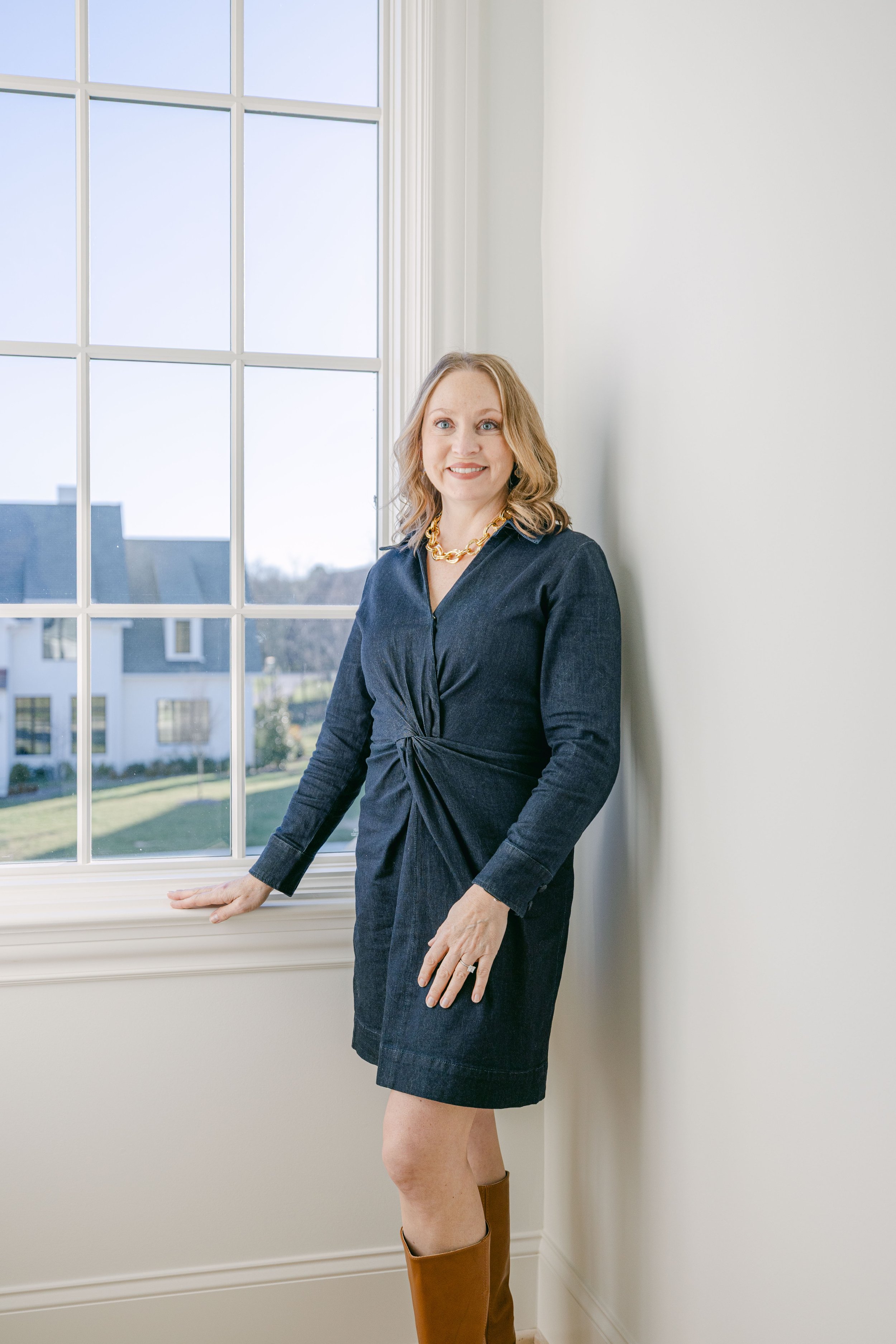 A woman wearing a dark dress and brown knee-high boots standing near a window, with a smile on her face inside a bright room.