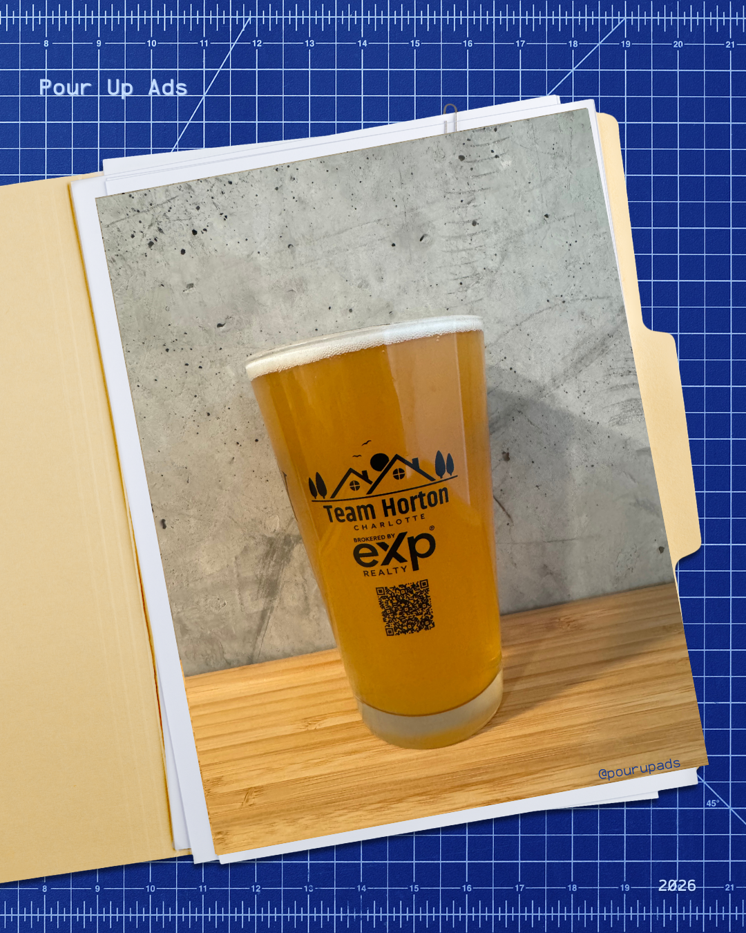 A printed photograph of a tall glass of beer with a foam head, placed on a wooden surface against a concrete wall background. The beer glass features a logo for Team Horton Charlotte, brokered by exp Realty, with a QR code below.