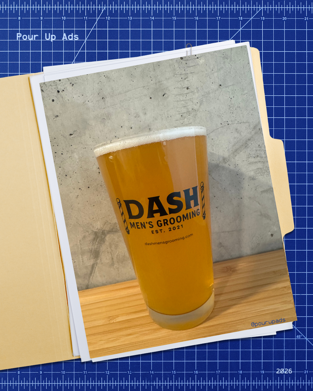 A pint glass of beer on a wooden surface with a concrete wall background, displaying the logo 'DASH Men's Grooming' and website.