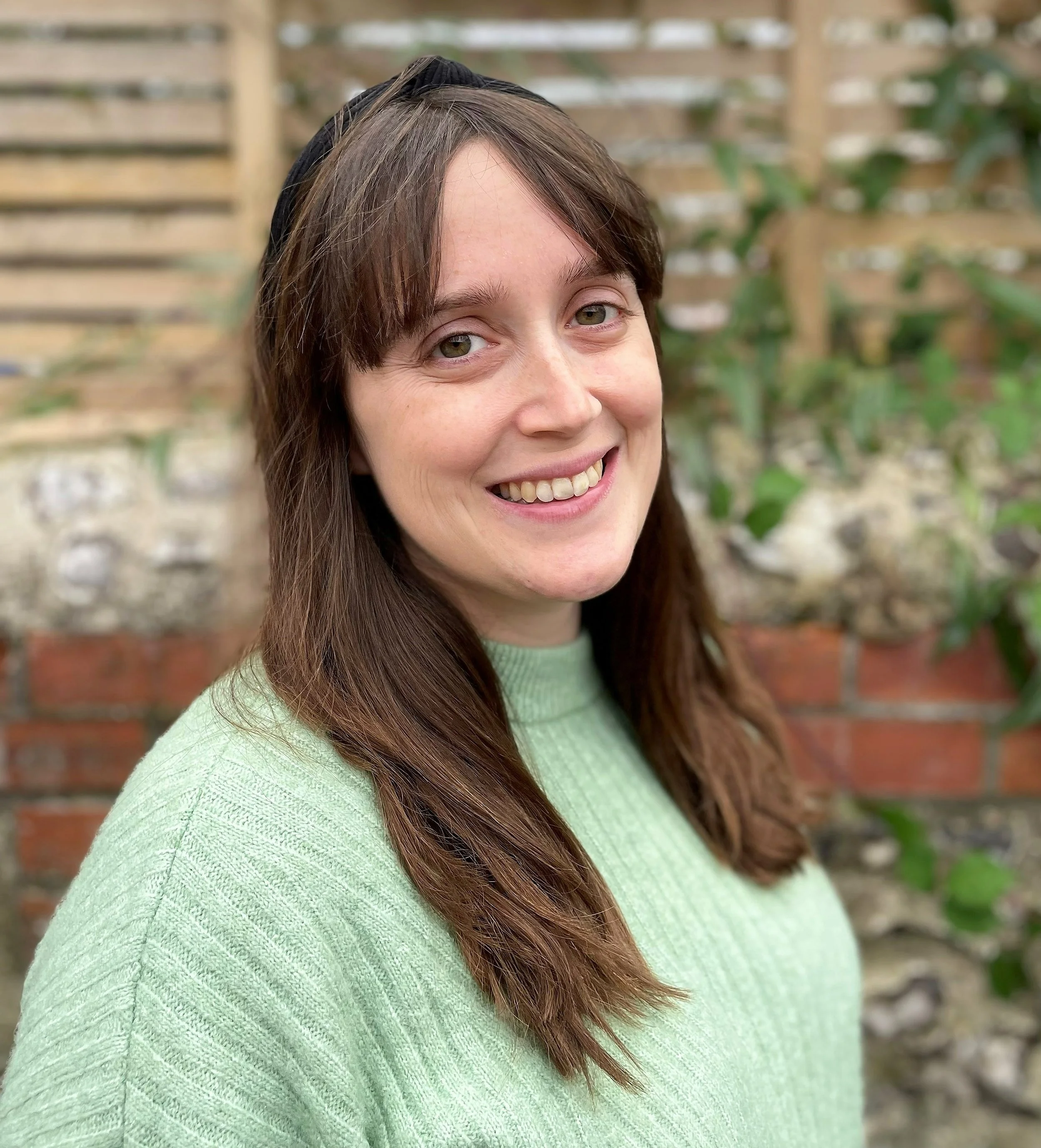 Close-up of a smiling woman with long brown hair, wearing a light green sweater and a black headband, standing outdoors in front of a brick wall and a wooden trellis with green plants.