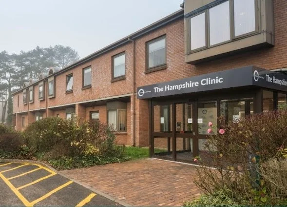 Exterior view of The Hampshire Clinic, a brick multi-story building with a black awning over the entrance, and a brick sidewalk leading to the door. There are bushes and a parking lot with yellow markings in front.