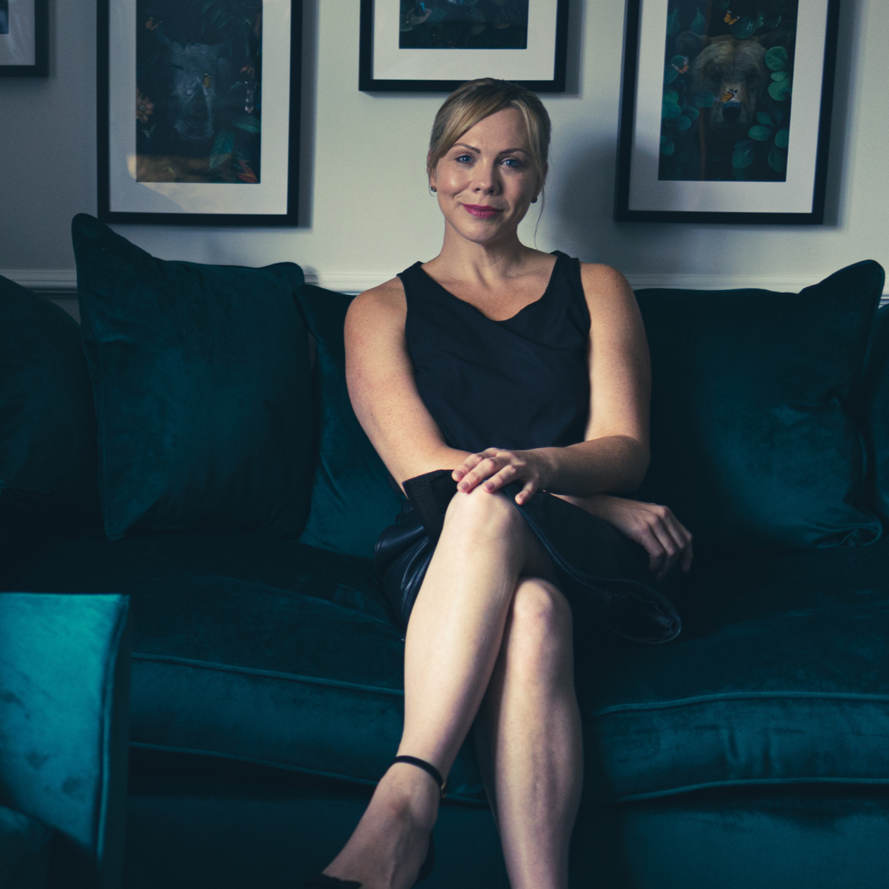 A woman sitting on a dark teal sofa with arm crossed, wearing a black sleeveless dress and black heels, smiling at the camera, with framed artwork on the wall behind her.