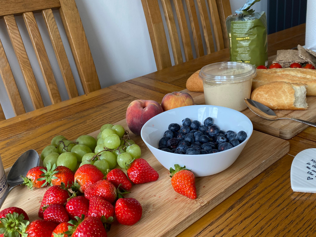 A wooden table with fresh strawberries, green grapes, blueberries, peaches, a bowl of dip or cream, a sliced loaf of bread, a small baguette, cherry tomatoes, a packet of chips, and a note with writing on it.
