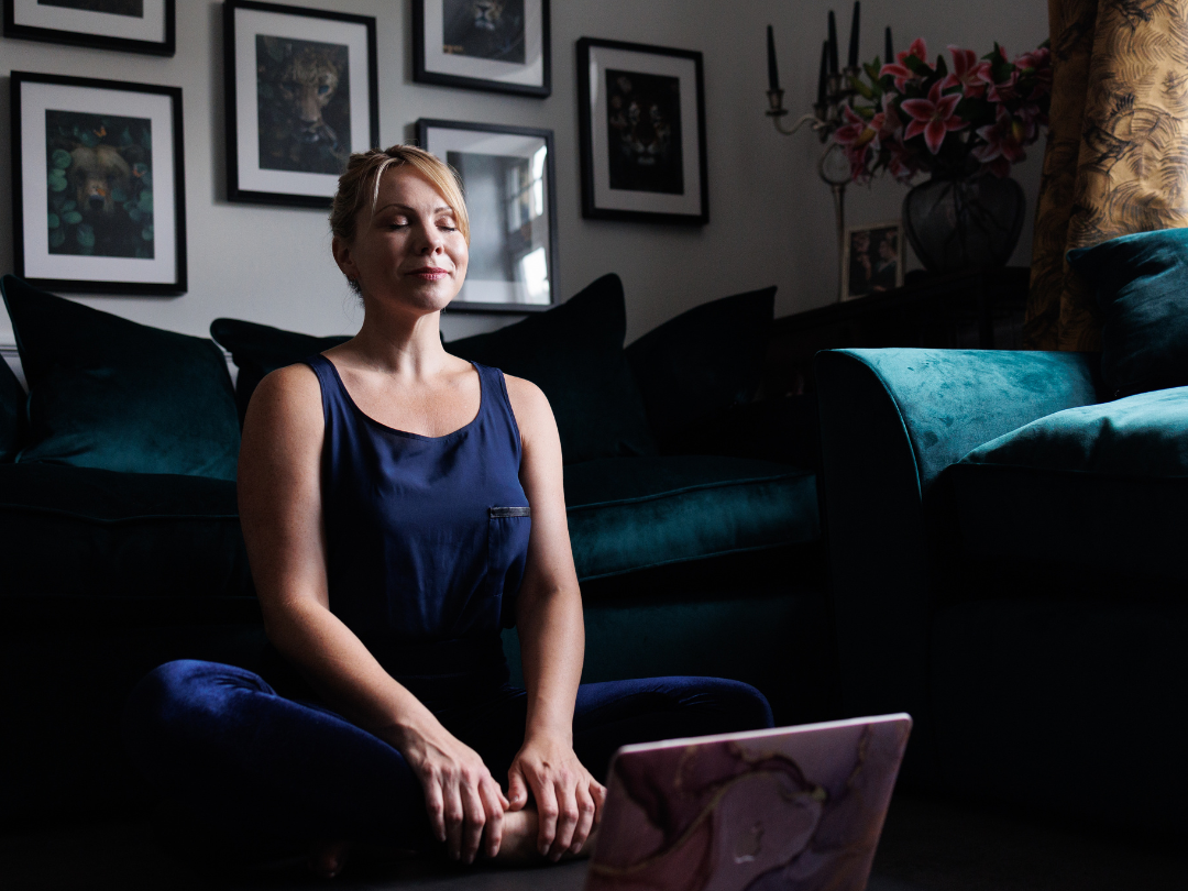 Claire Anstey sitting on the floor with eyes closed, meditating, in a living room with framed artwork, a dark green sofa, and a laptop in front of her.