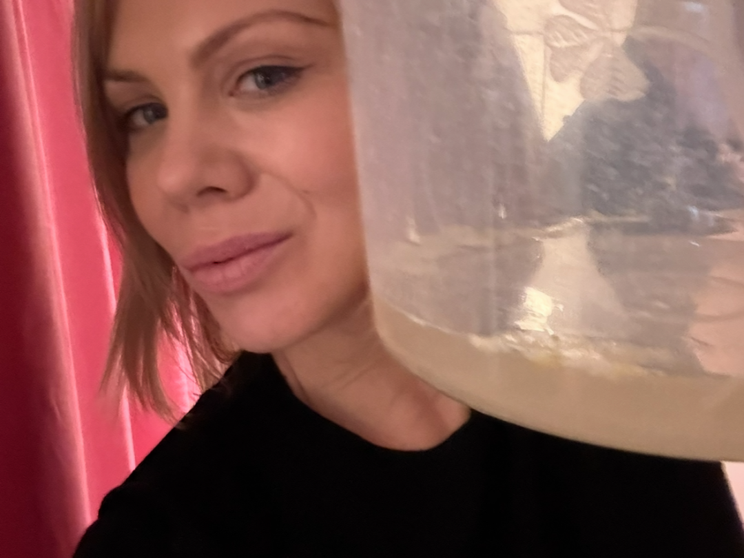A woman with short hair holding a clear plastic container close to her face, with a pink curtain in the background.