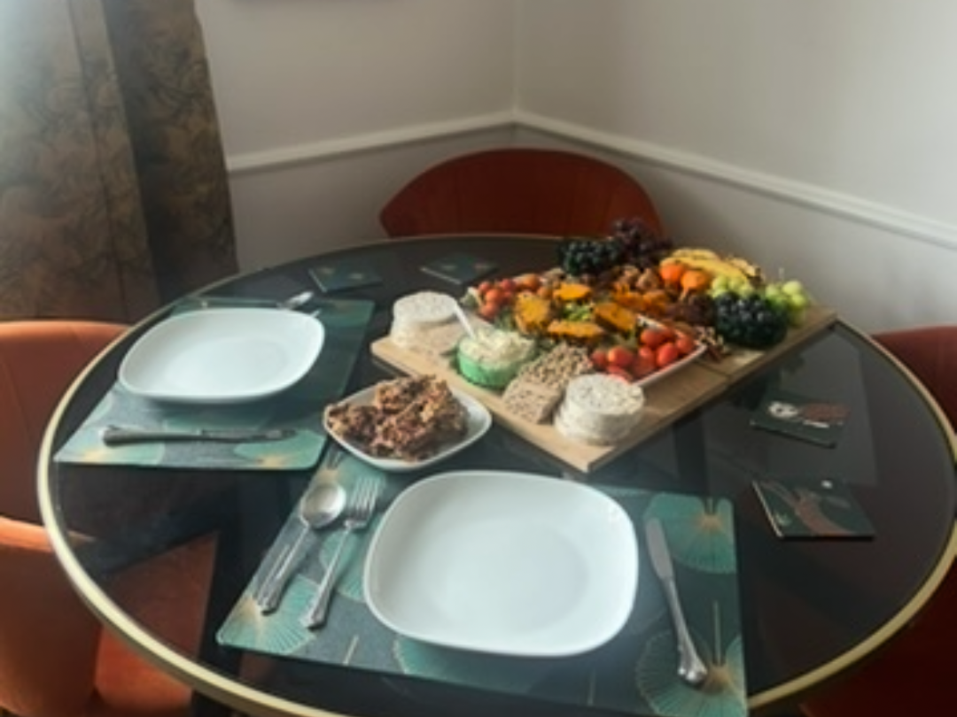 Round dining table set for three, with empty square white plates, spoons, and forks on patterned placemats. In the center, there's a wooden tray with an assortment of fruits, cheeses, and crackers, along with a small bowl of spread or dip. Three chai