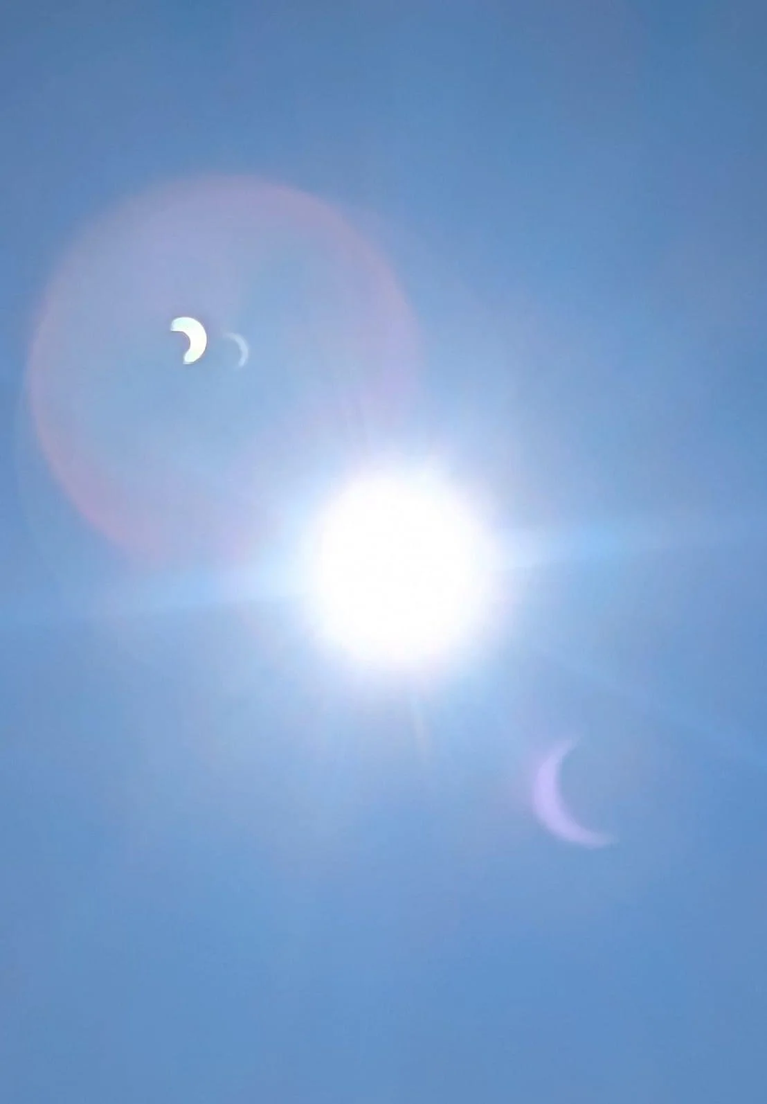 Bright sun in a clear blue sky with a visible crescent moon and lens flares.
