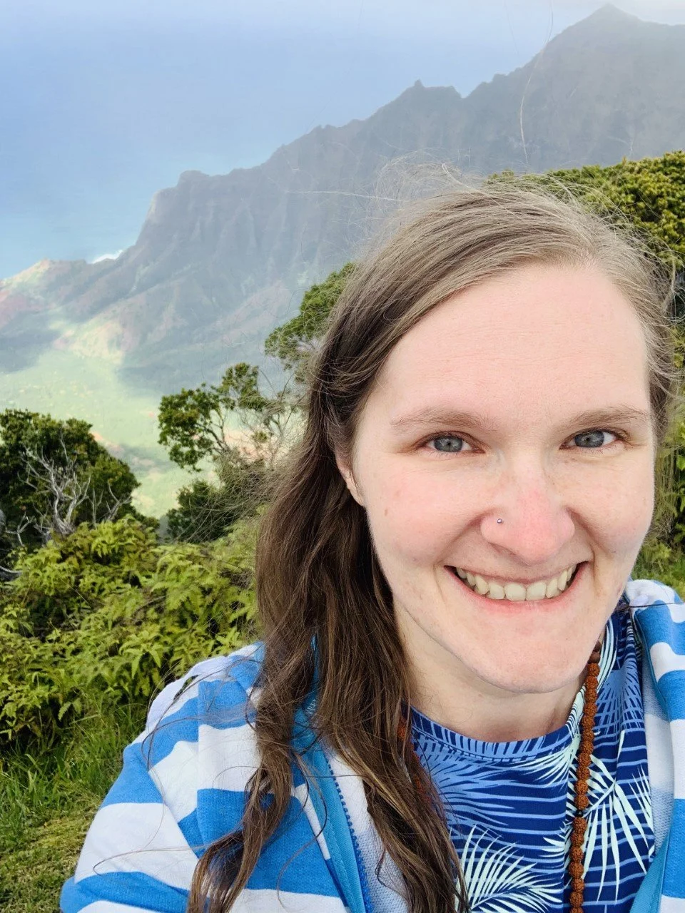 A smiling woman with long brown hair and a nose piercing outdoors with green mountains and lush foliage in the background.