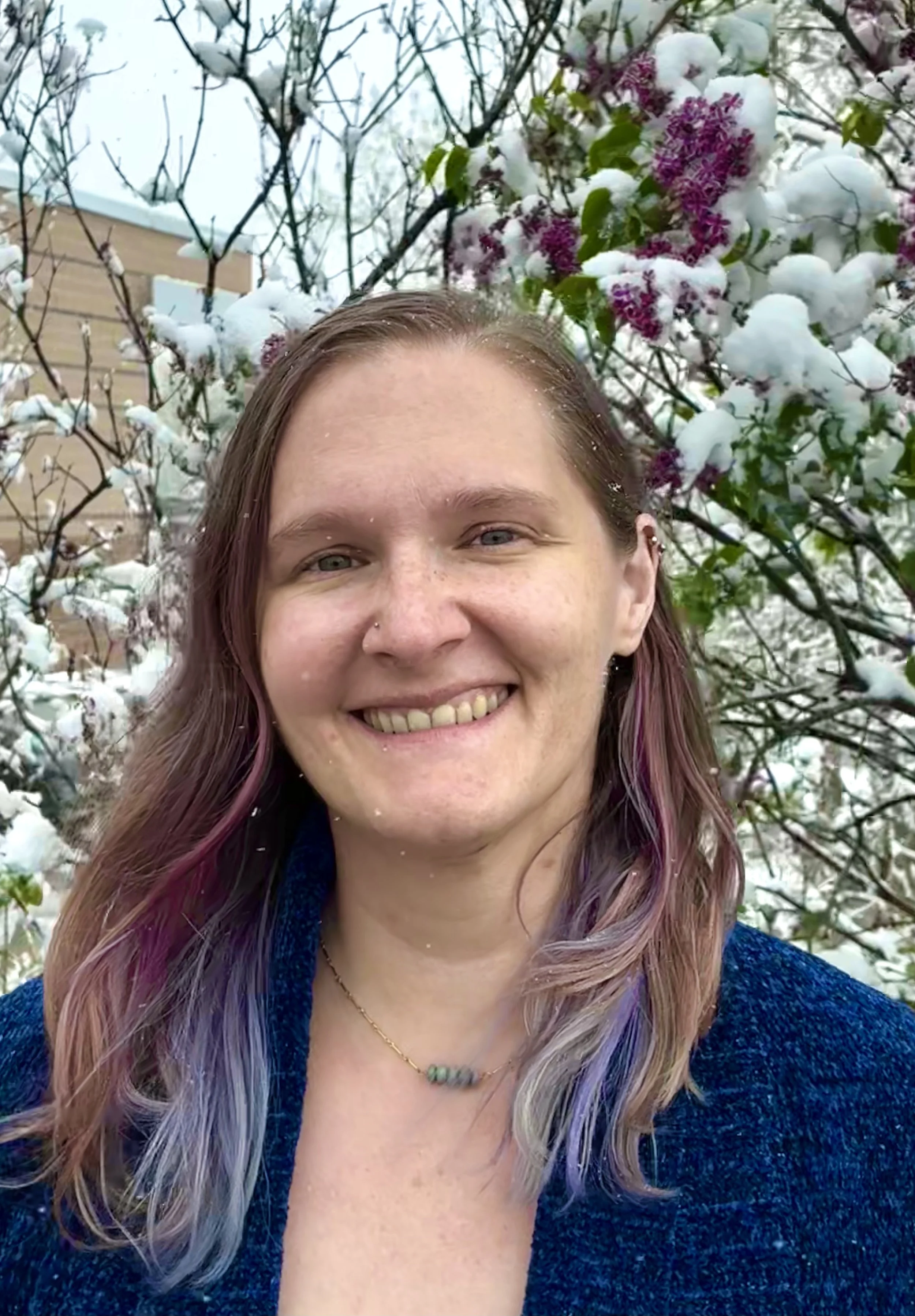 Close-up of a woman smiling outdoors, with snow-covered branches and pink and white blossoms in the background.