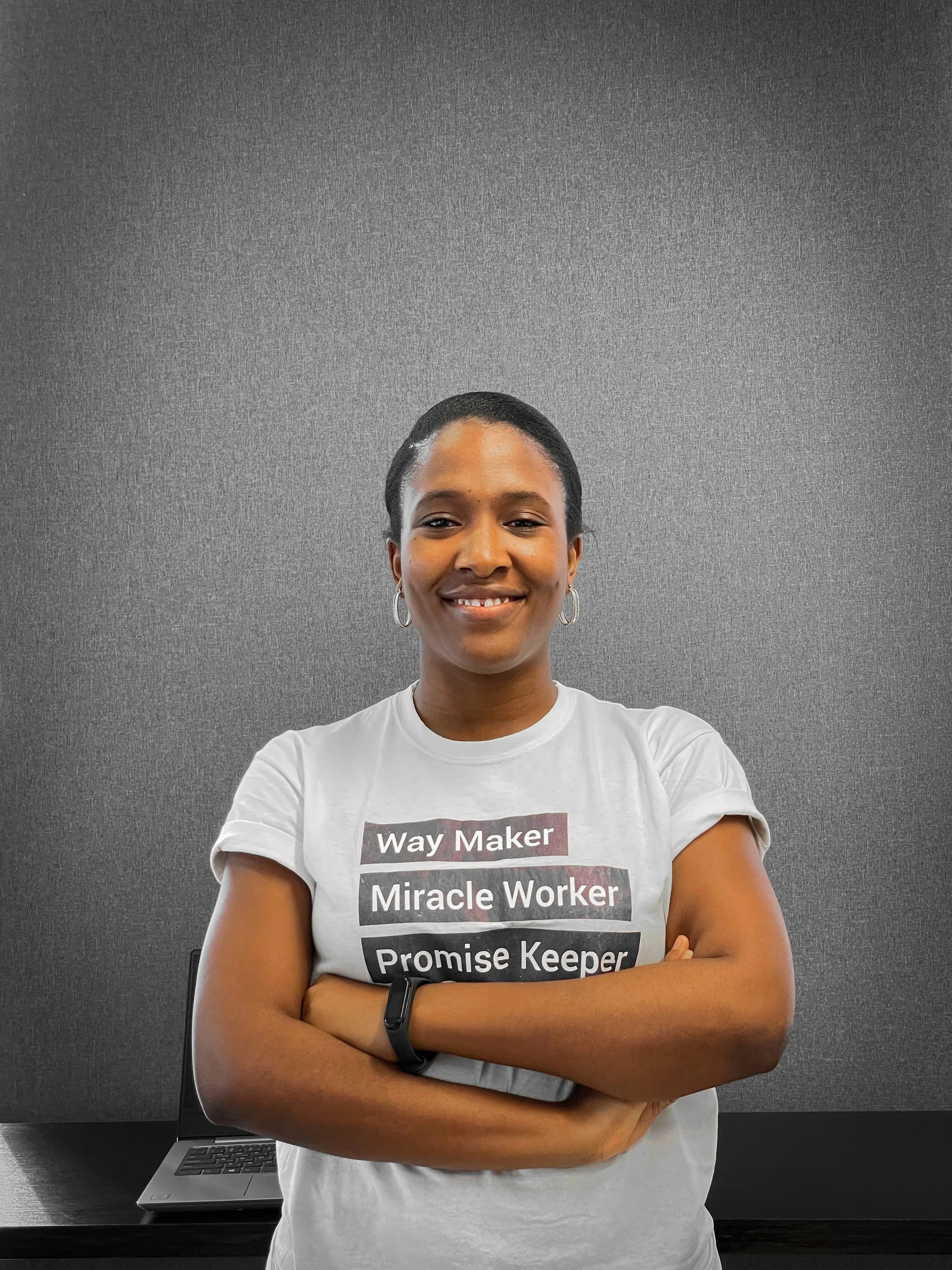 A woman with short black hair and hoop earrings smiling with her arms crossed, standing in front of a gray wall and a laptop on a black desk.