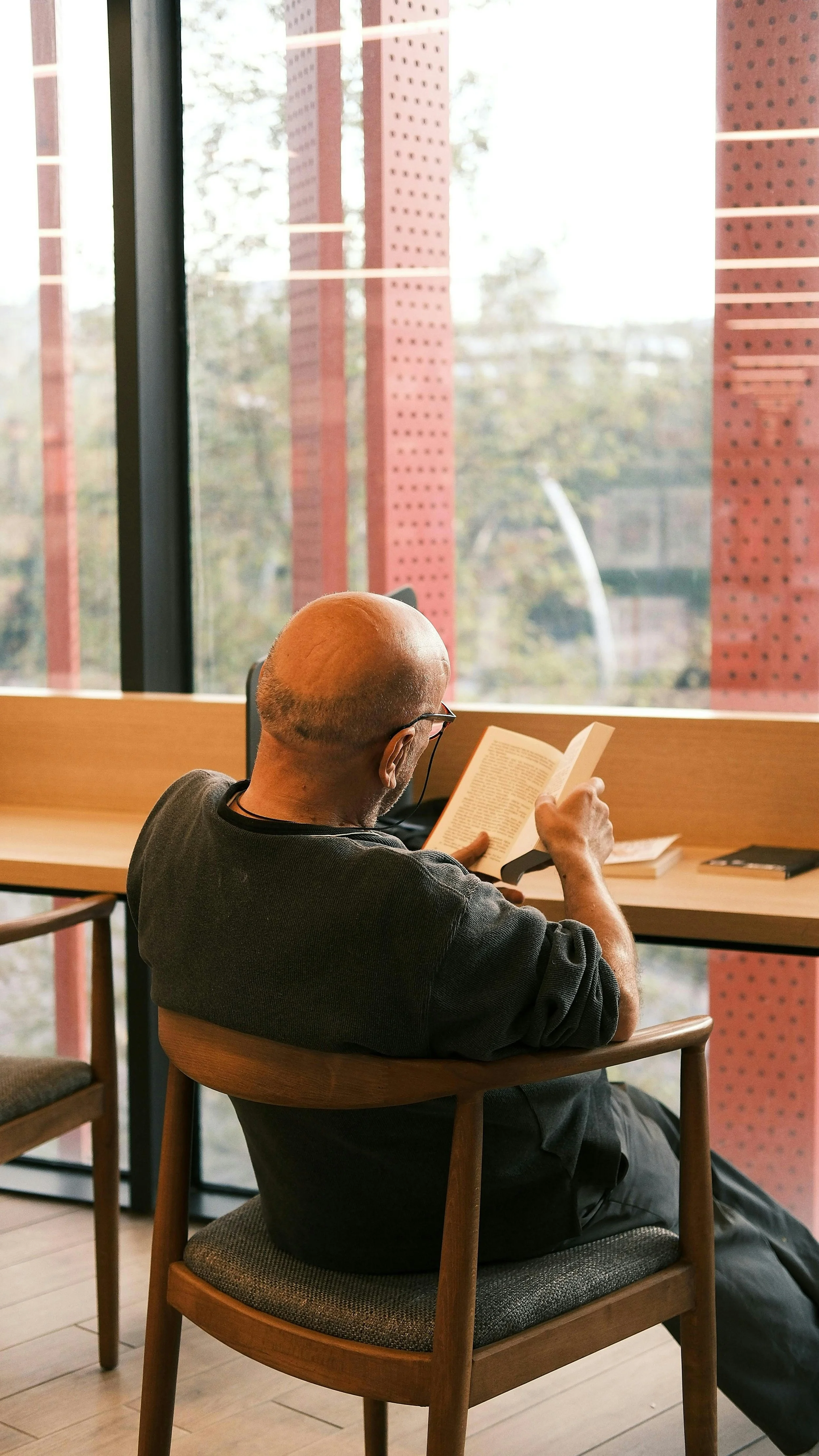 A man with a bald head and glasses sits in a wooden chair, reading a book at a cafe near a large window with a view of trees and red structural supports outside.