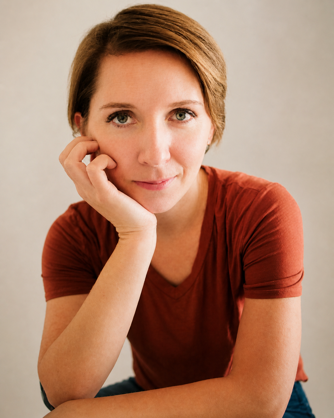 A woman with short brown hair and green eyes gazing at the camera, resting her face on her left hand with her elbow supported on a surface, wearing a rust-colored shirt.