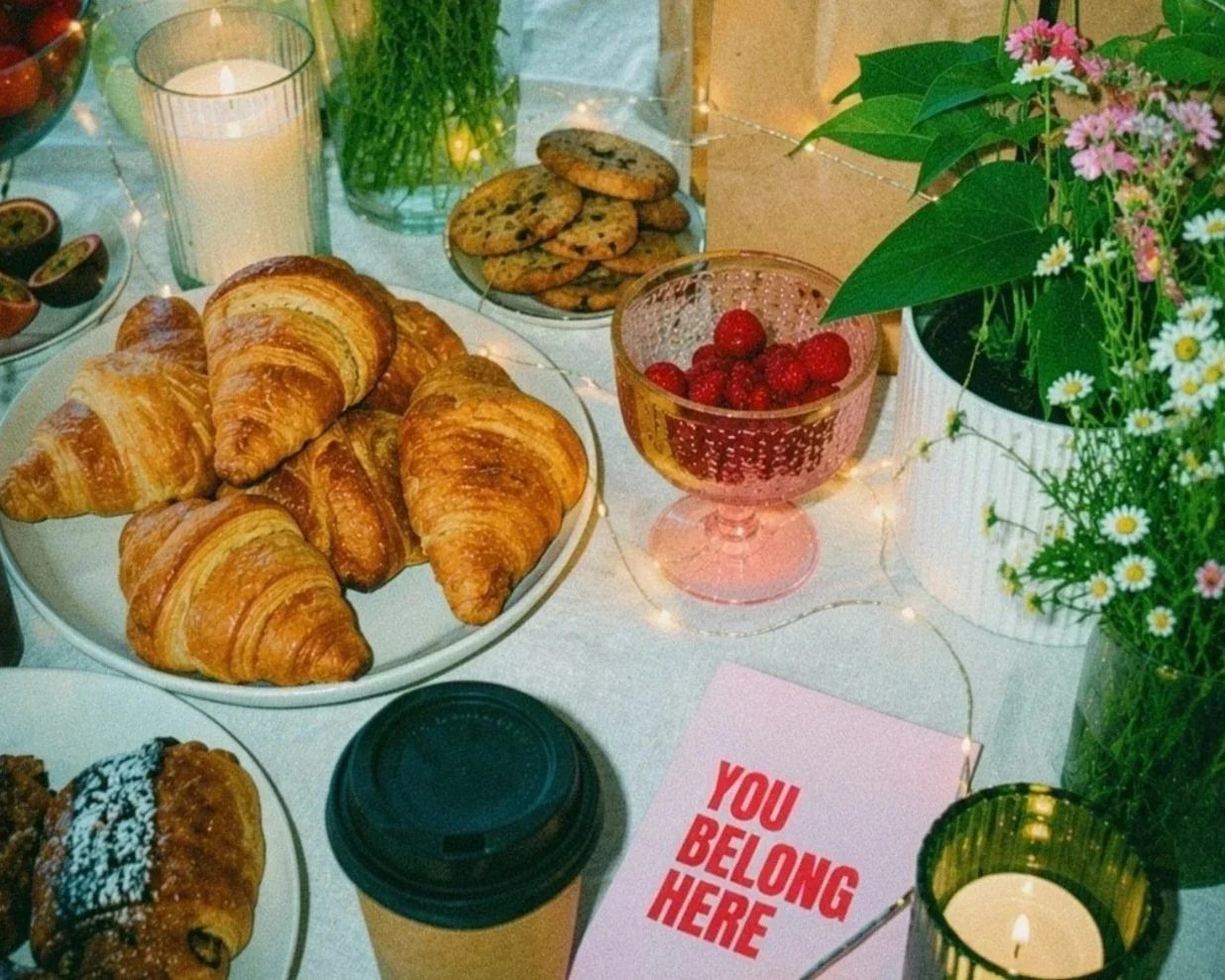 A table with croissants, cookies, a bowl of strawberries, and a variety of flowers in vases. There is a candle, a coffee cup, and a sign that reads 'You Belong Here'.