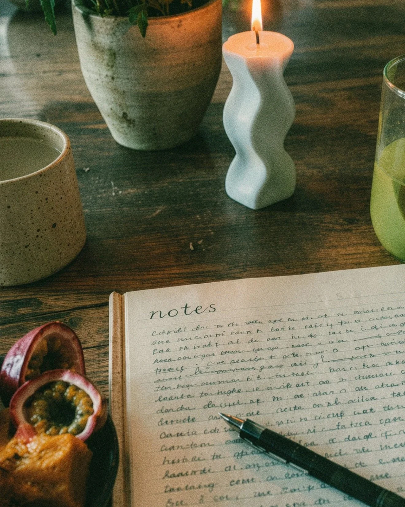 A top view of a wooden table with a potted plant, a lit white candle in a wavy holder, two cups of beverage, a partially visible notebook with handwritten notes and a pen, and halved passion fruits.